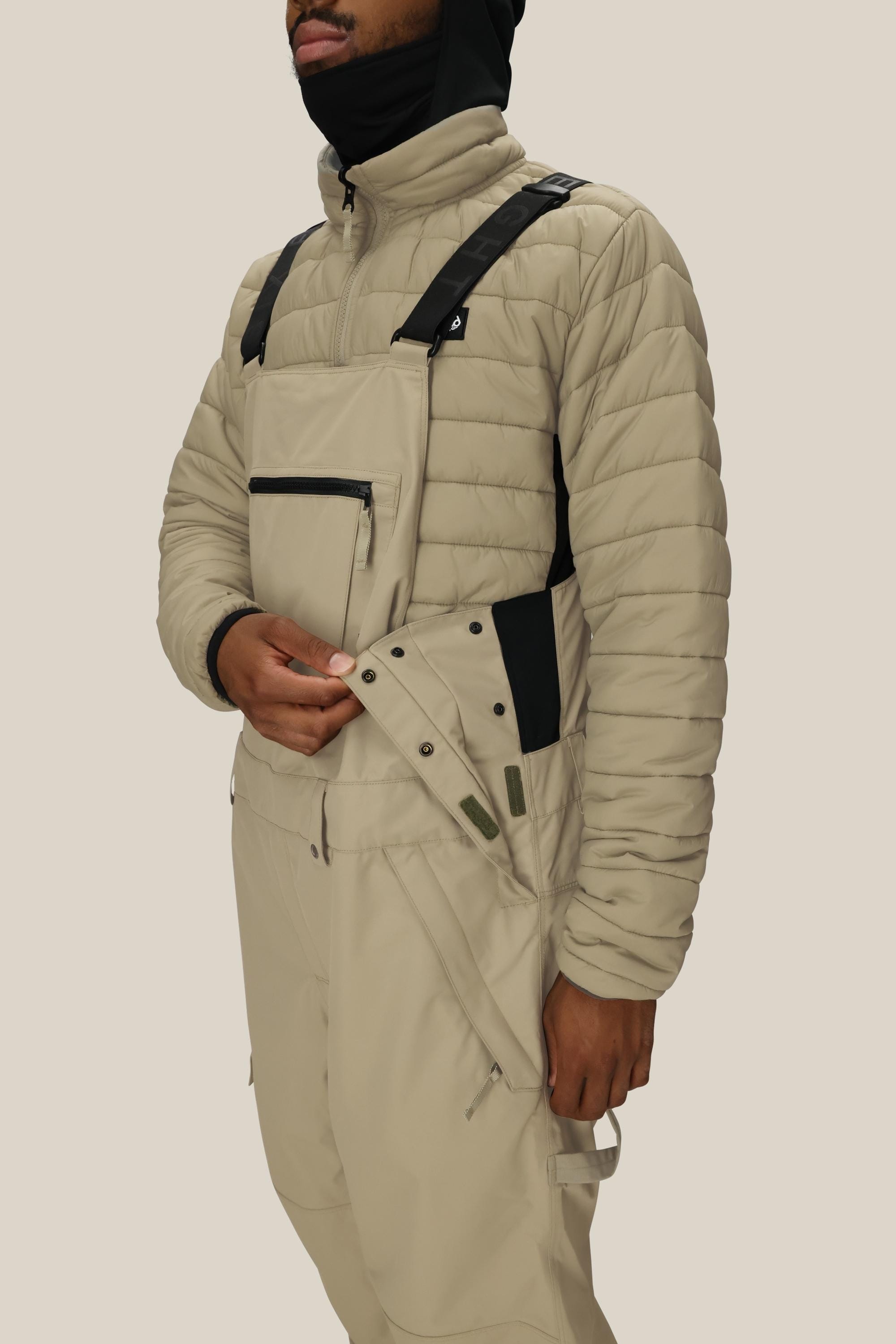 A person, face partly covered by a black hood, fastens the side buttons of beige 686 Men's 20K Hot Lap™ Shell Bib overalls with black straps, paired with a beige quilted jacket.
