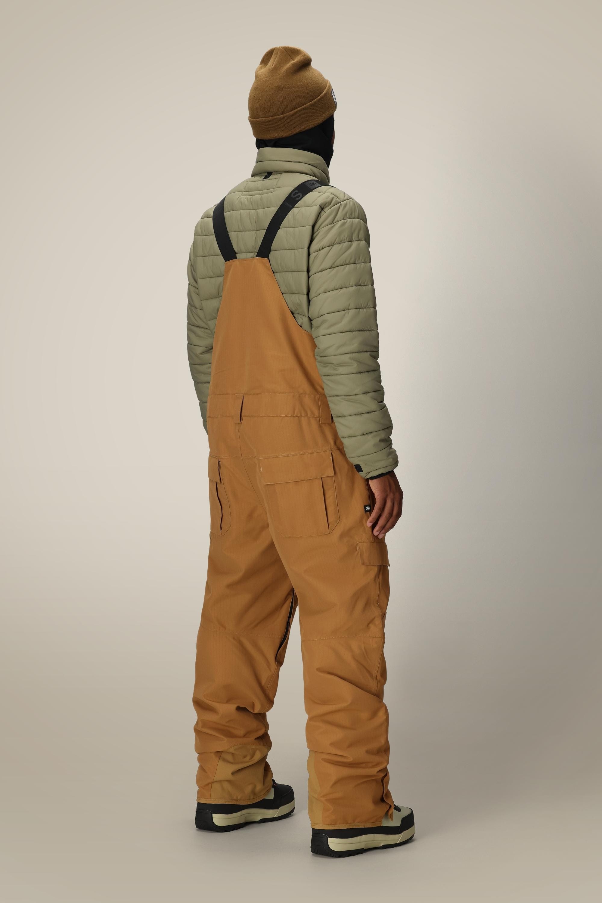 A person stands facing away in a tan beanie, olive quilted jacket, 686 Men's Hot Lap™ Insulated Bib by 686 with black straps, and black boots against a neutral background.
