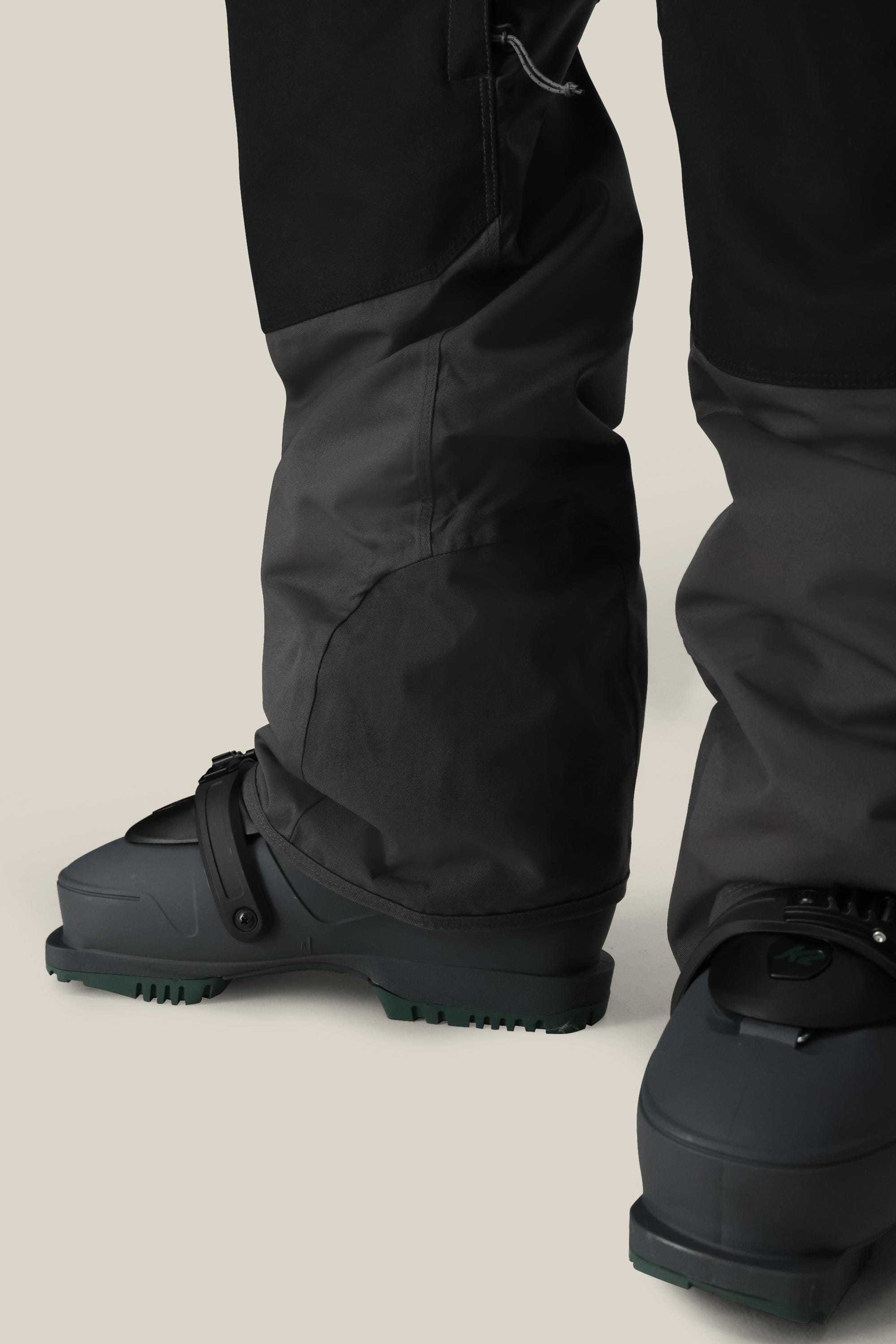 Close-up of a person wearing 686 Men's Hot Lap™ Insulated Bib snowboard pants in dark, waterproof polyester-nylon-spandex blend, partially covering their dark gray boots, standing on a neutral beige background. Brand: 686.