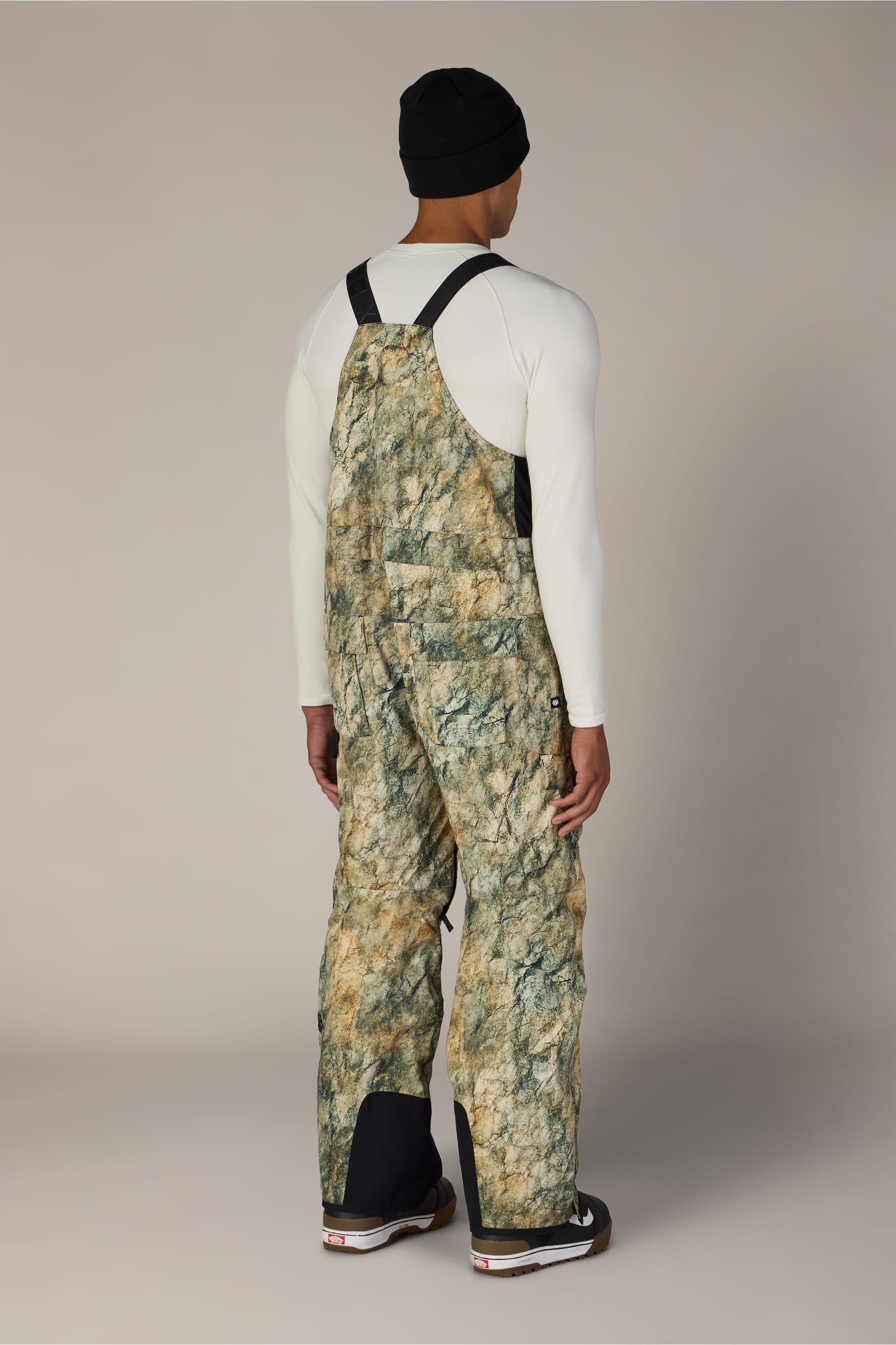 A person stands facing away, wearing a black beanie, a fitted off-white long-sleeve shirt, and the 686 Men’s Hot Lap™ Insulated Bib by 686 in green-brown camouflage with black straps and details, paired with dark shoes against a plain background.