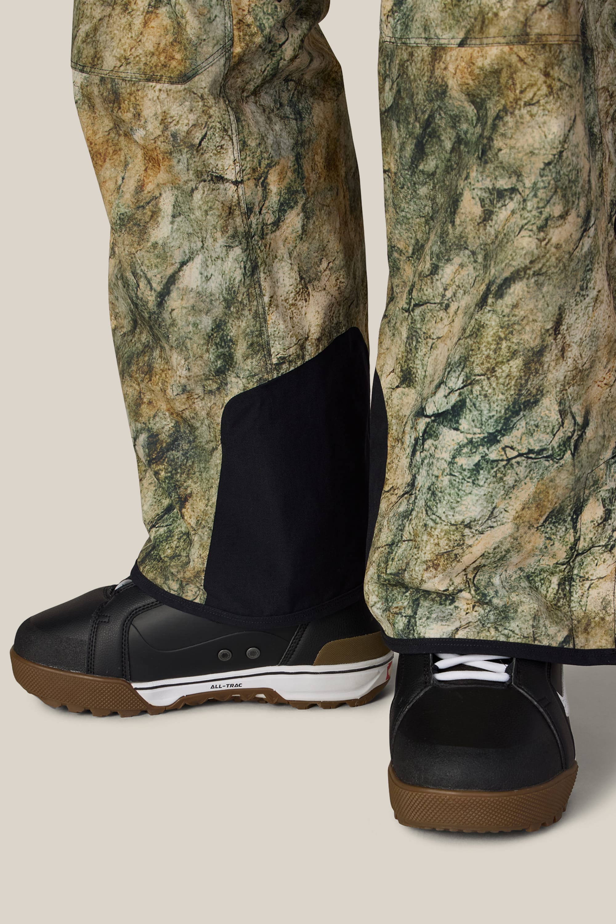 Close-up of a person wearing the 686 Men’s Hot Lap™ Insulated Bib in a camouflage pattern, paired with black sneakers featuring white soles and brown accents, standing on a neutral background.