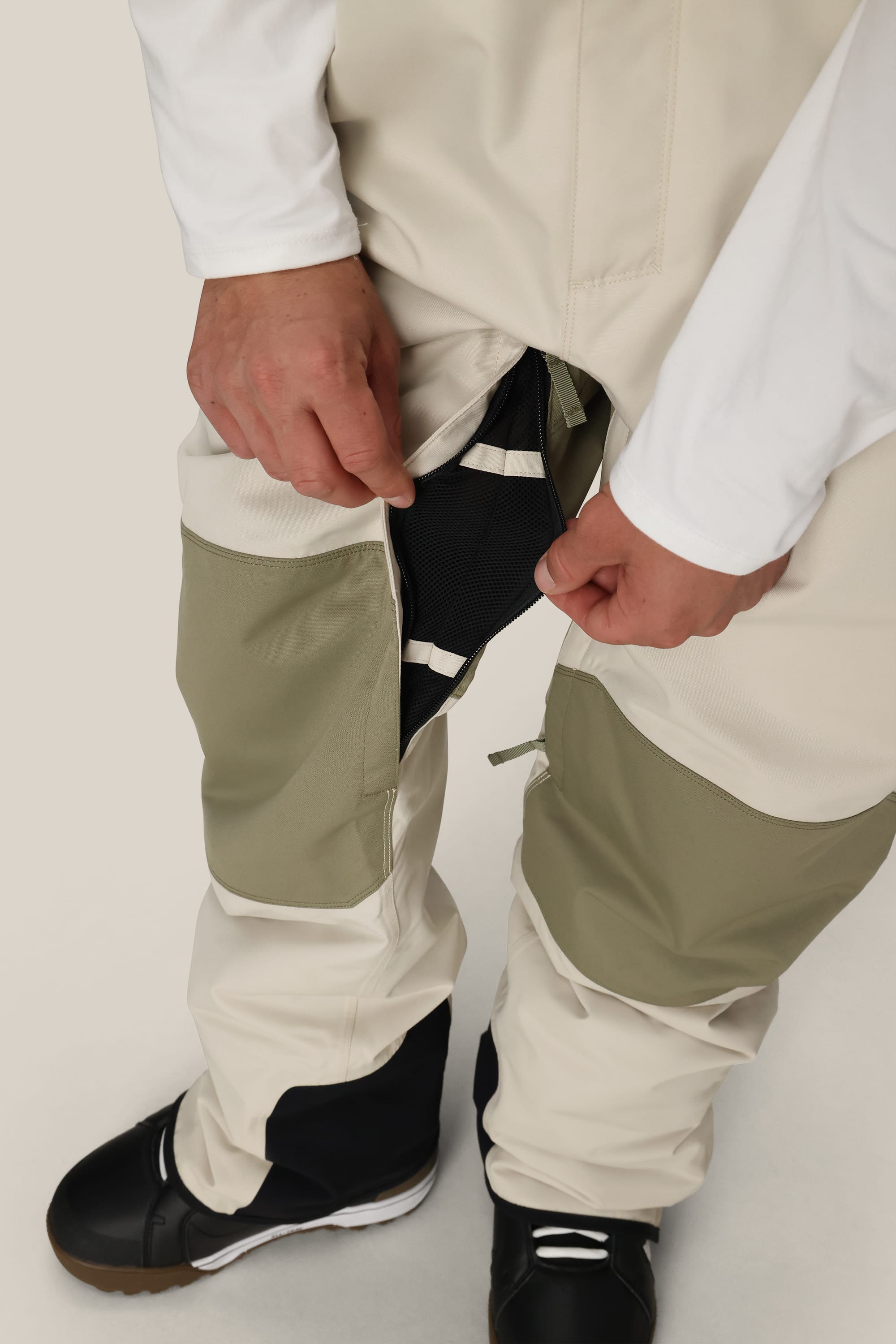 Wearing the 686 Men's Hot Lap™ Insulated Bib in two-tone beige and olive green, a person opens a thigh zipper to reveal black mesh ventilation. They're also dressed in a white long-sleeve shirt and black shoes.