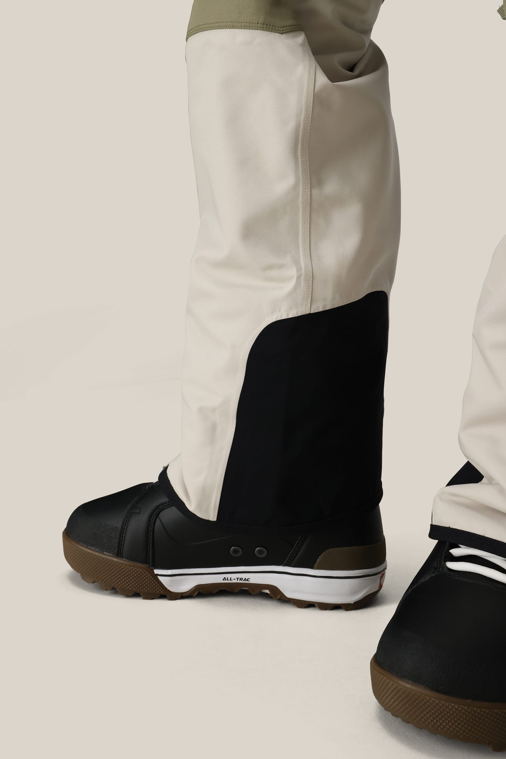 A close-up of a person wearing the 686 Men's Hot Lap™ Insulated Bib by 686, paired with black and brown winter boots, standing on a light beige background.