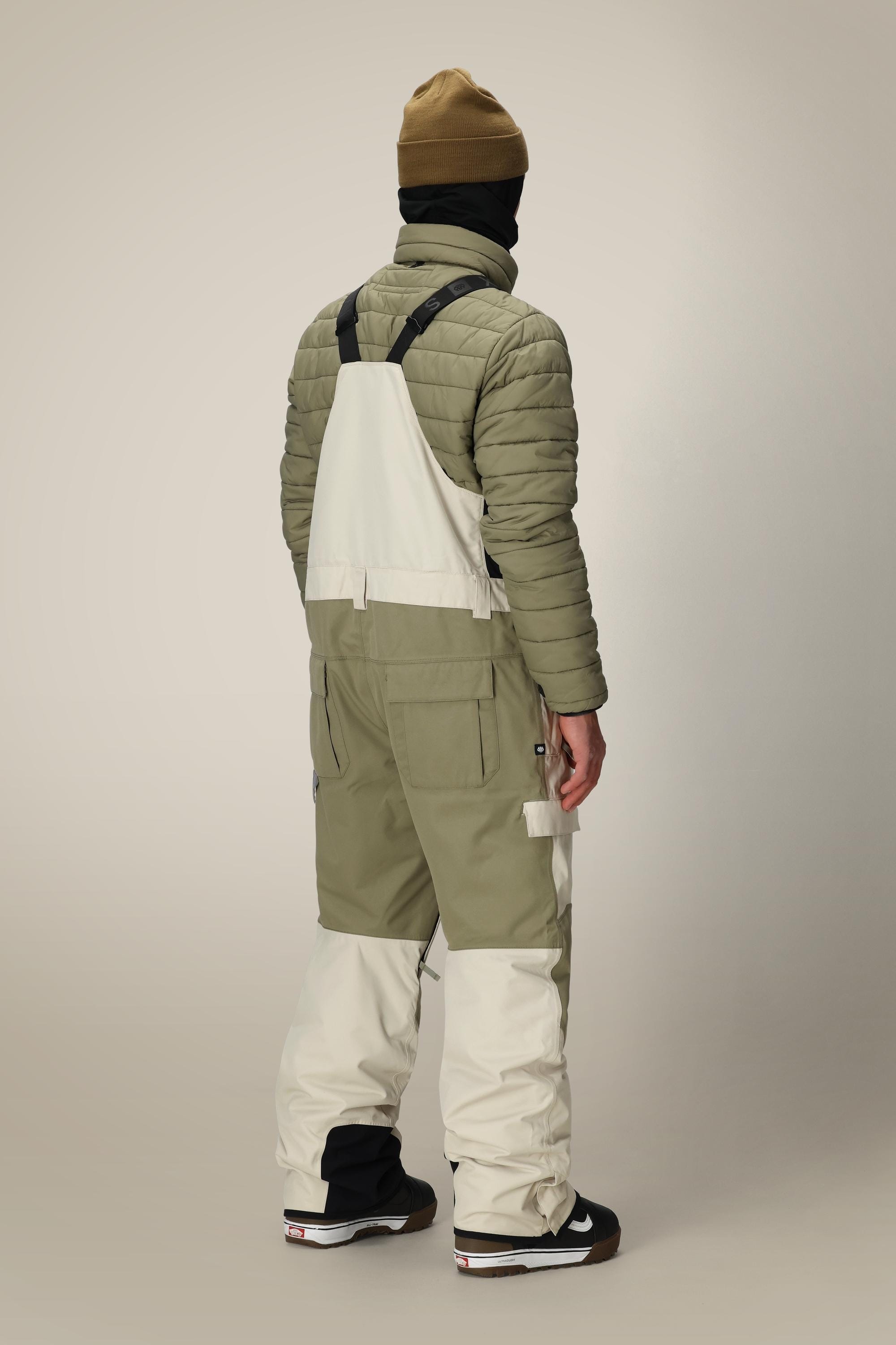 A person stands facing away, wearing a tan beanie, black neck gaiter, olive green quilted jacket, and 686 Men's Hot Lap™ Insulated Bib by 686. Black and white sneakers pop against the plain light background.
