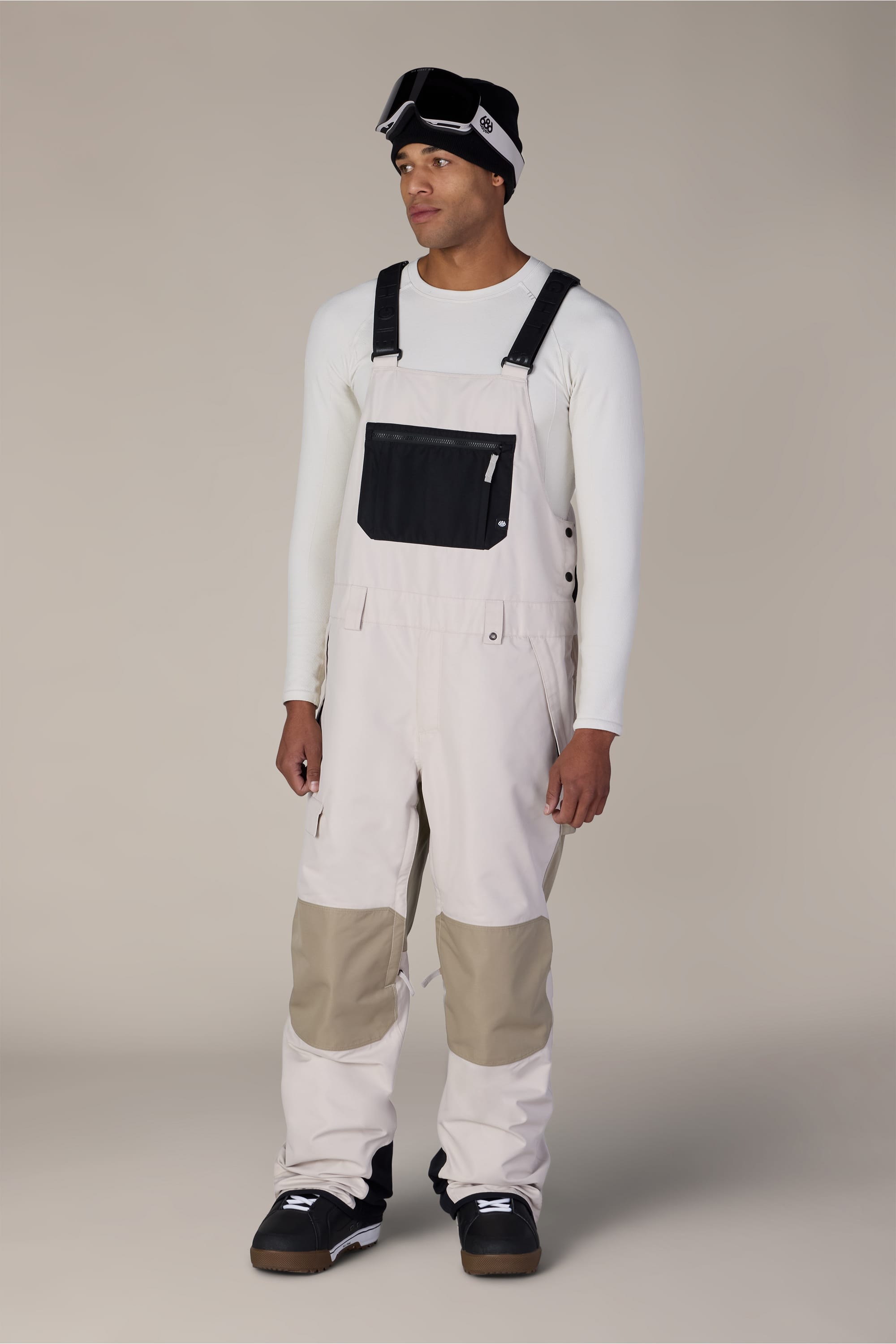 A person wearing the 686 Men’s Hot Lap™ Insulated Bib in a light color with a black chest pocket, paired with a white long-sleeve shirt, black and white shoes, ski goggles on their forehead, and a white beanie, standing against a plain background.