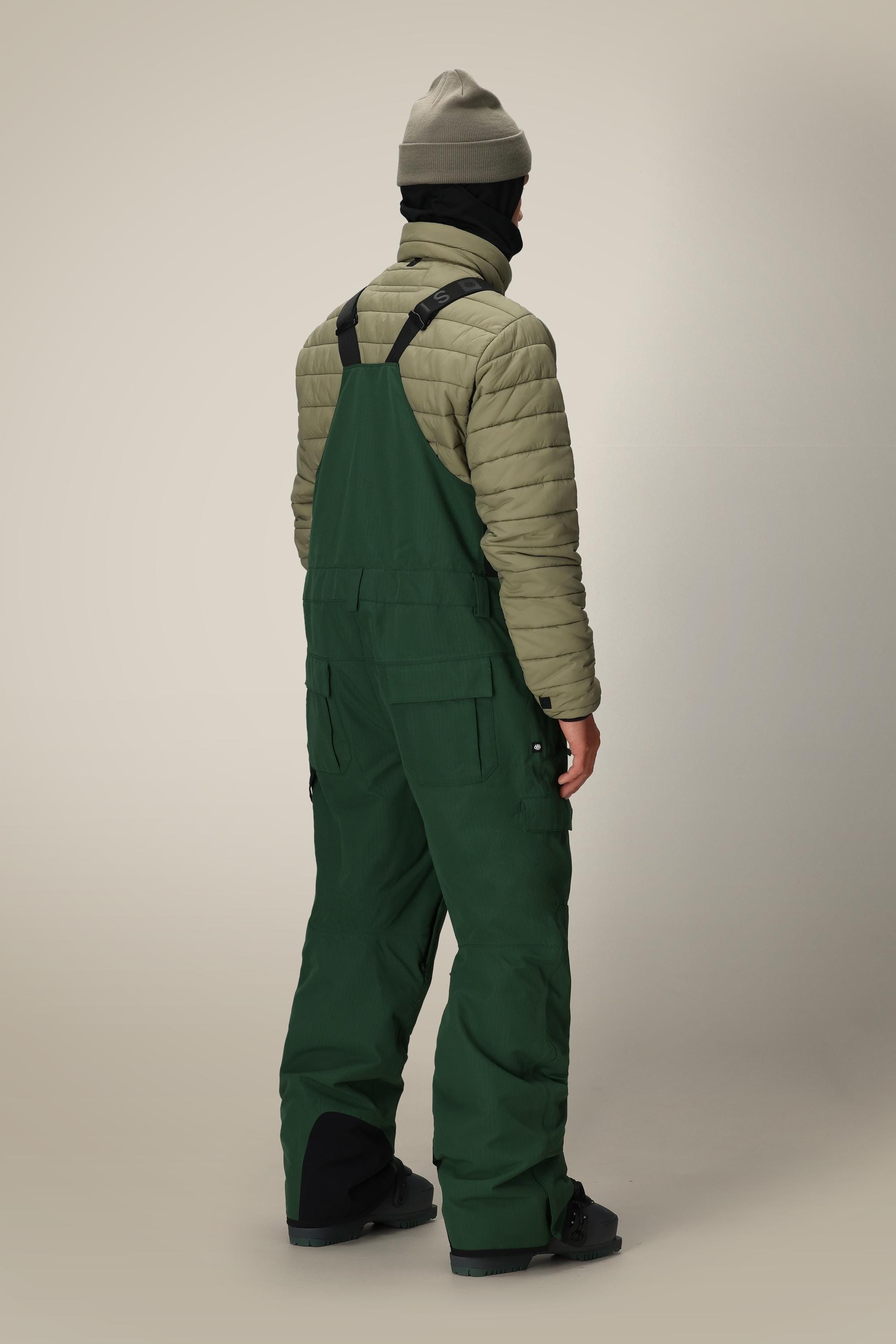A person stands facing away, wearing a green beanie, a tan quilted jacket, black boots, and dark green 686 Men's Hot Lap™ Insulated Bib overalls with black straps against a neutral background.