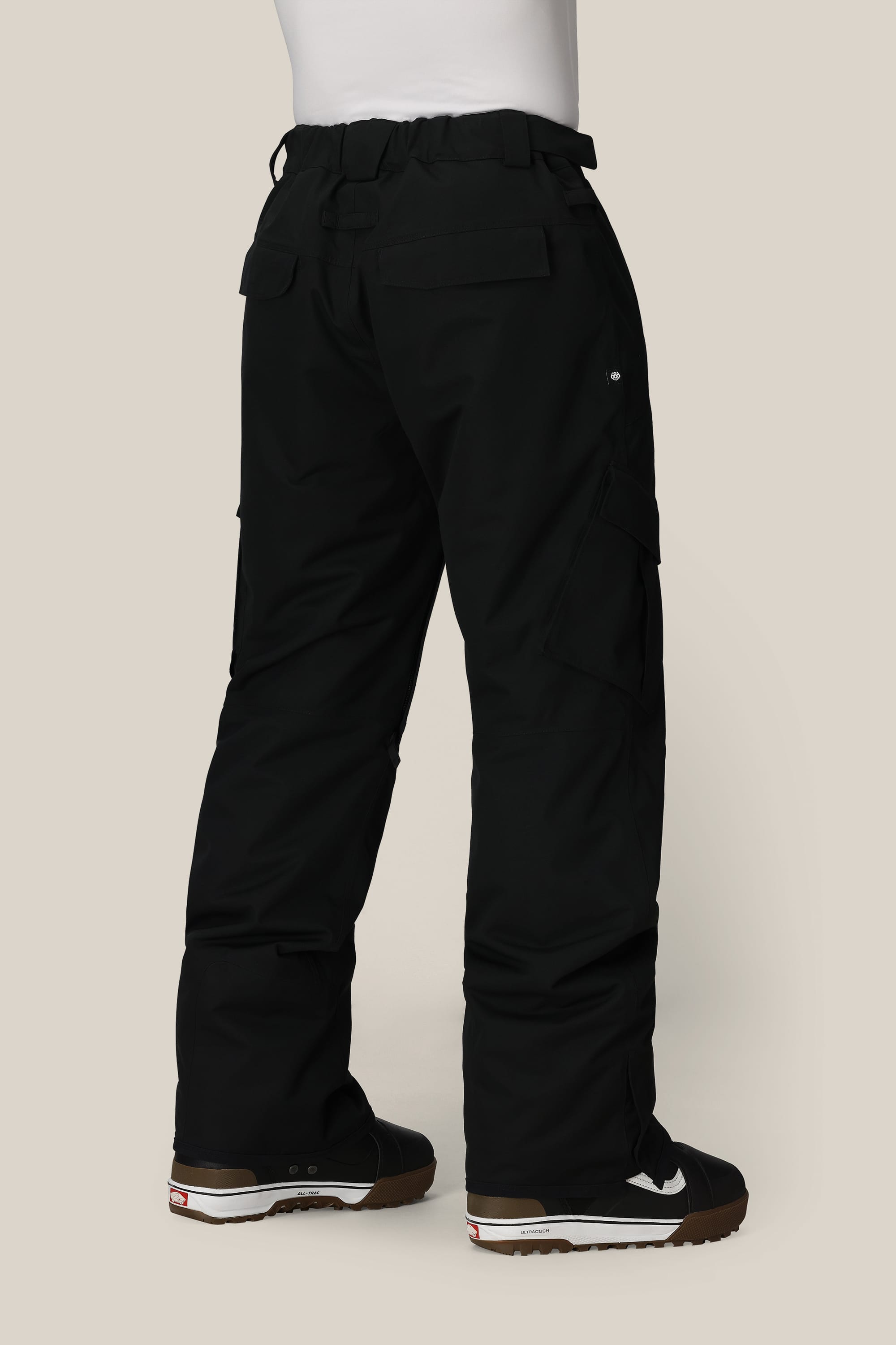 A person wearing 686 Men's Infinity™ Insulated Cargo Pants and black sneakers with white accents stands on a light beige background, shown from the back.