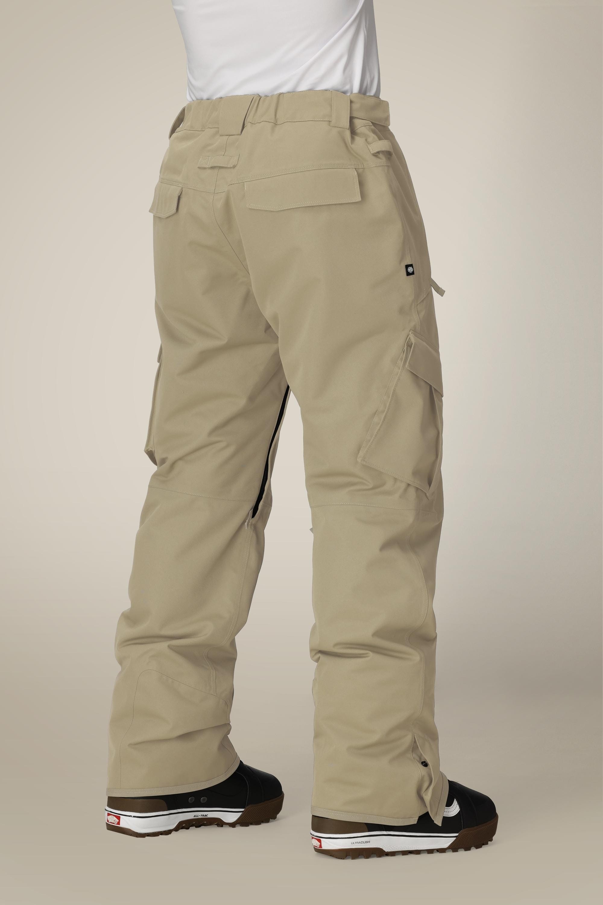 A person wearing 686 Men's Infinity™ Insulated Cargo Pant in tan with black boots stands facing away, highlighting the back and side cargo pockets. The background is plain and neutral.