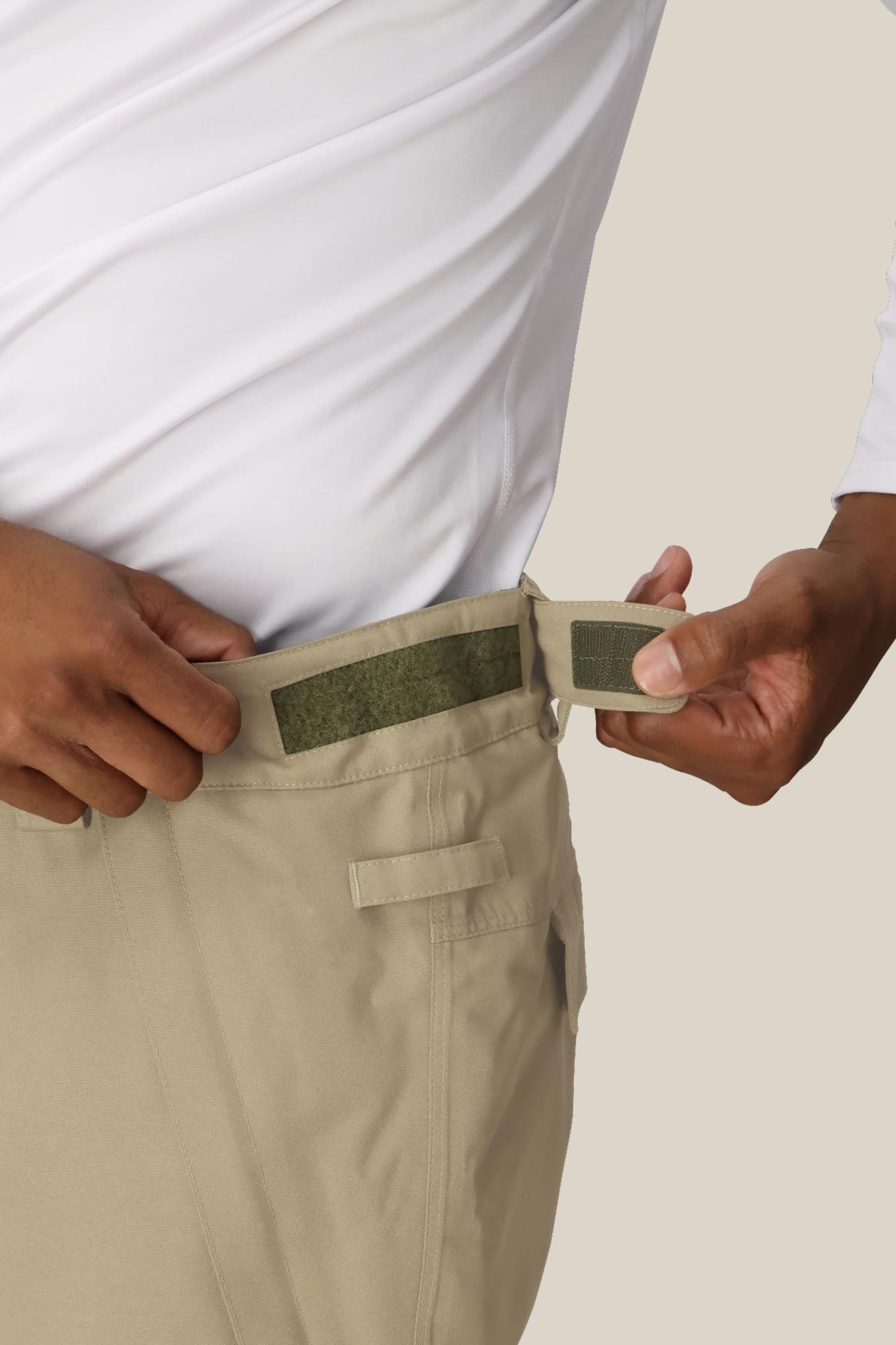A man wearing the 686 Men's Infinity™ Insulated Cargo Pant in beige and a white shirt adjusts the waistband using the Velcro strap on the side.