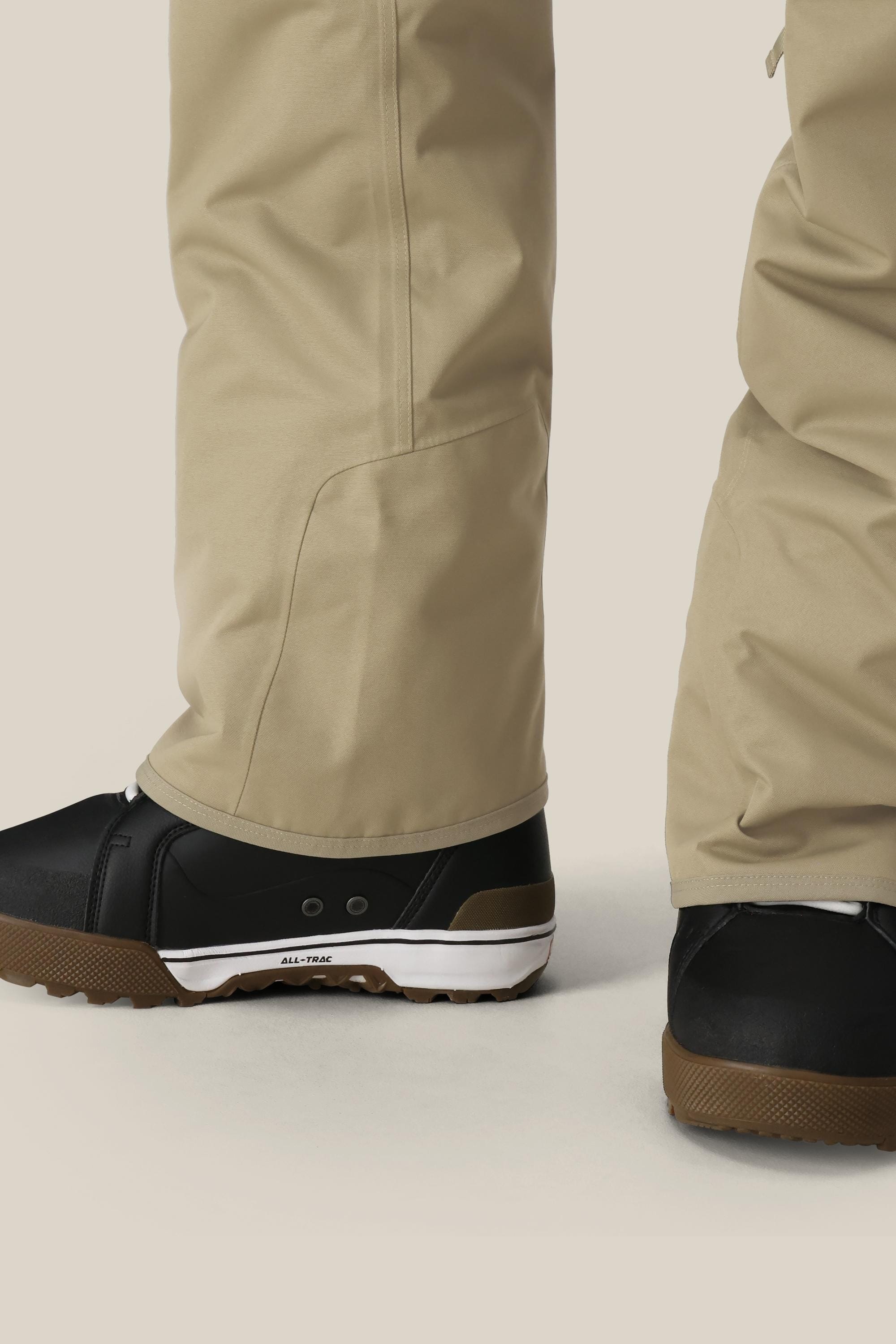 Two lower legs wear 686 Men's Infinity™ Insulated Cargo Pants in beige, paired with black sneakers featuring white soles, standing on a light surface. The focus is on the pants by 686 and the footwear.