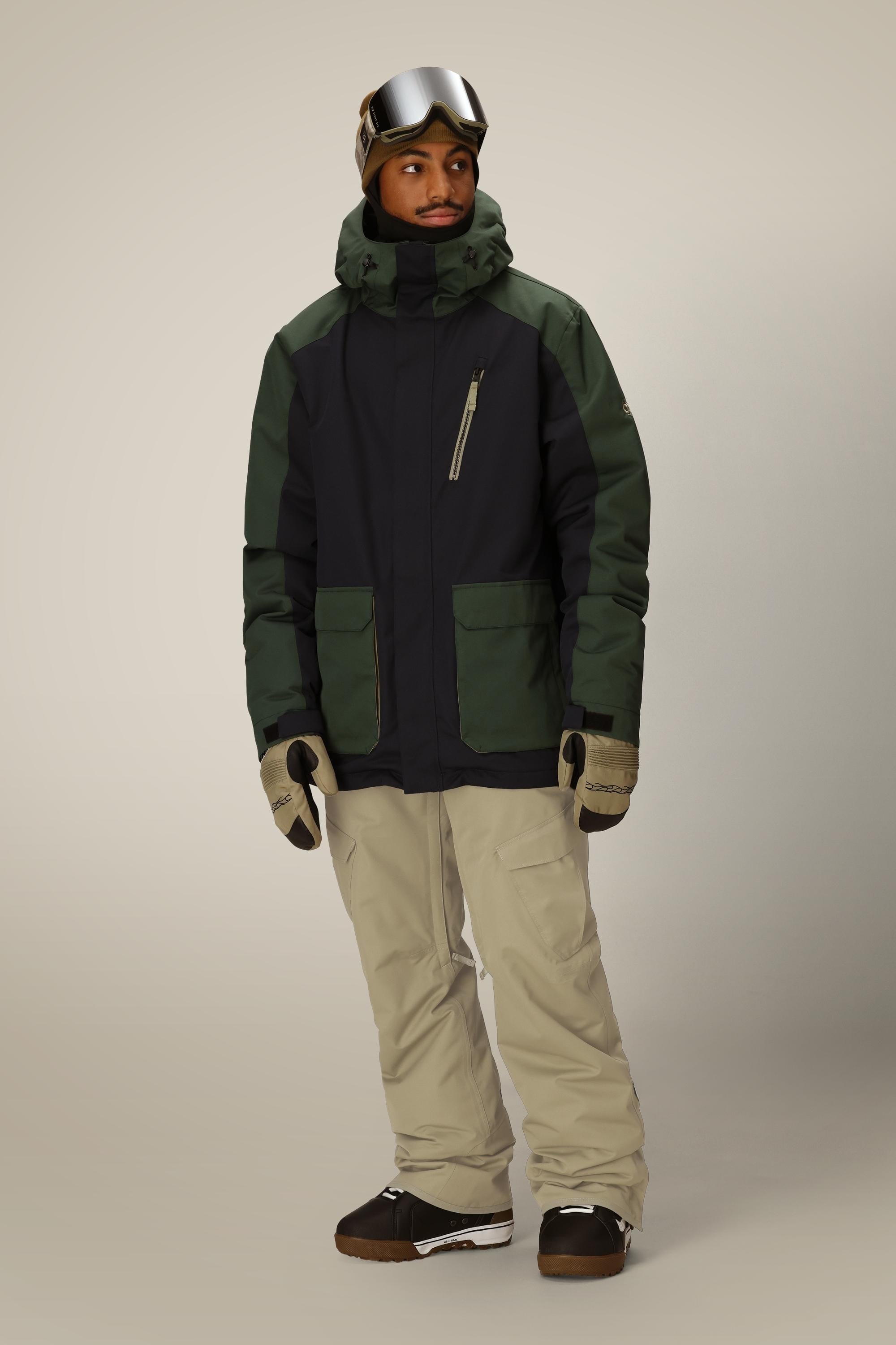 A person wearing a dark green and black winter jacket, tan gloves, brown snow boots, ski goggles, and 686 Men's Infinity™ Insulated Cargo Pant by 686 stands against a plain light background.