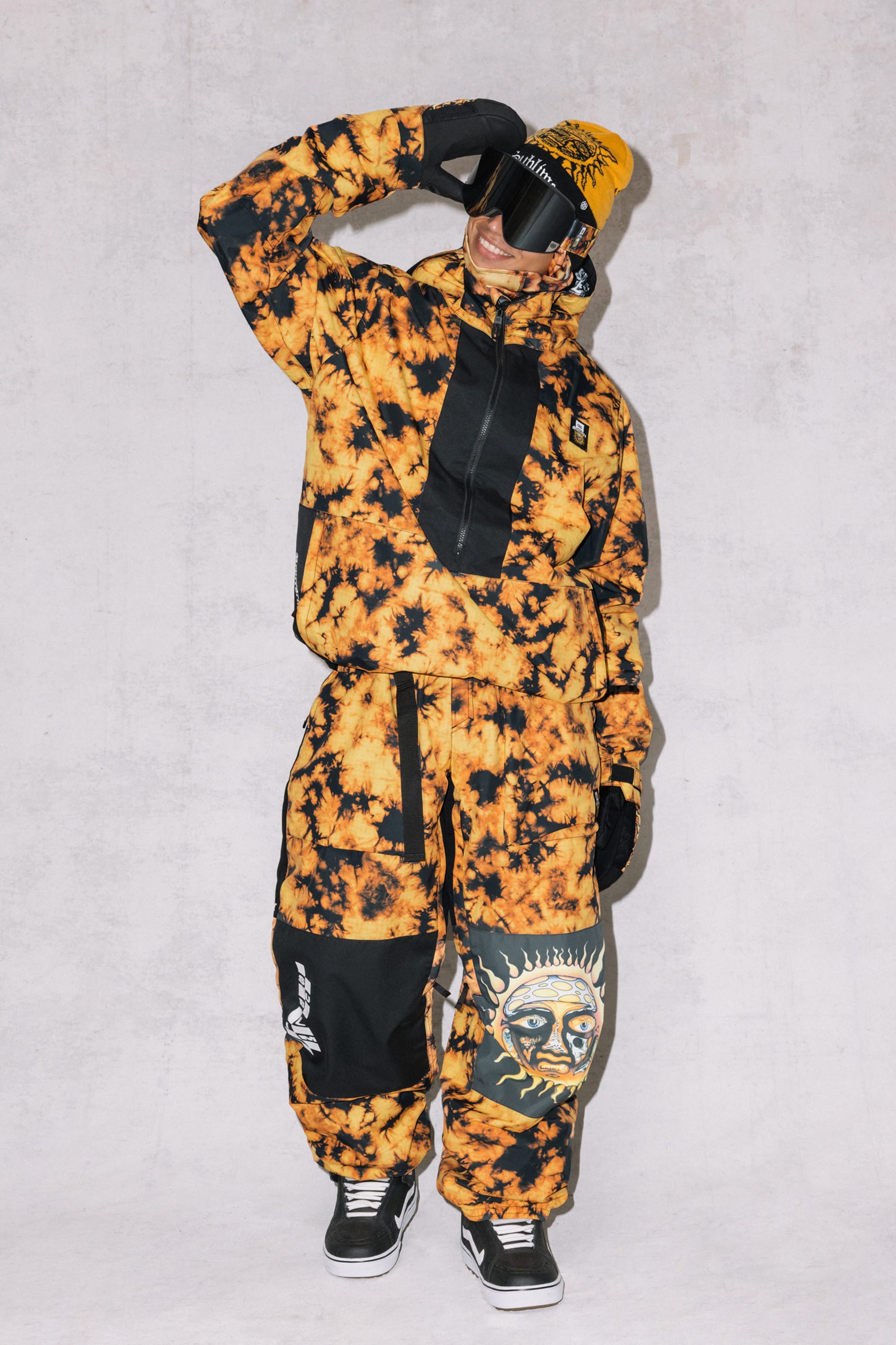 A person in a black and orange tie-dye outfit with sun and snake patches stands against a light background, wearing 686 Mens Sublime Shell Pants, a yellow beanie, black gloves, dark sunglasses, and black sneakers while posing with one hand saluting.