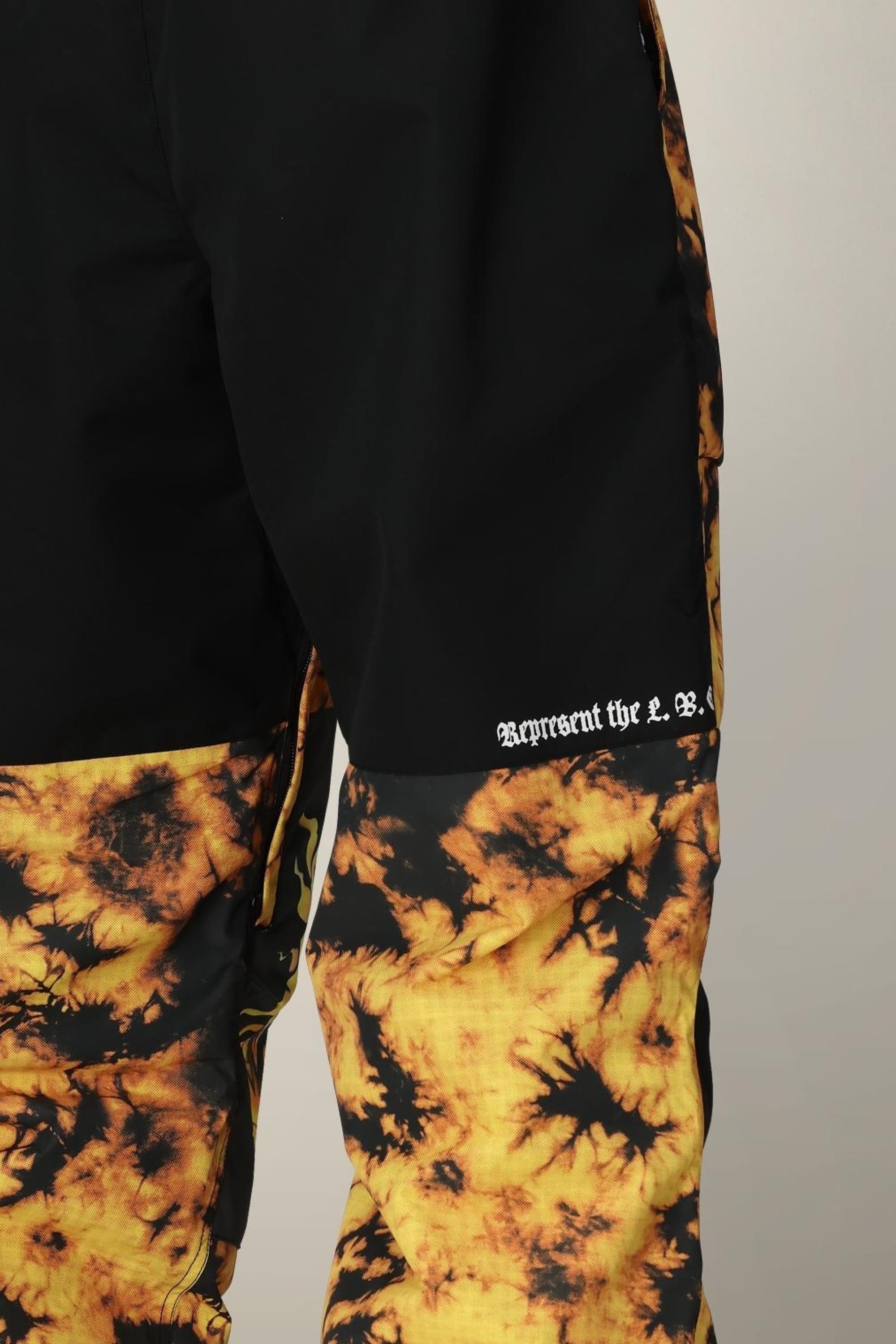 A close-up of the 686 Mens Sublime Shell Pants, featuring a black and yellow tie-dye design with Represent the L.B. in white Gothic-style text on the upper thigh.