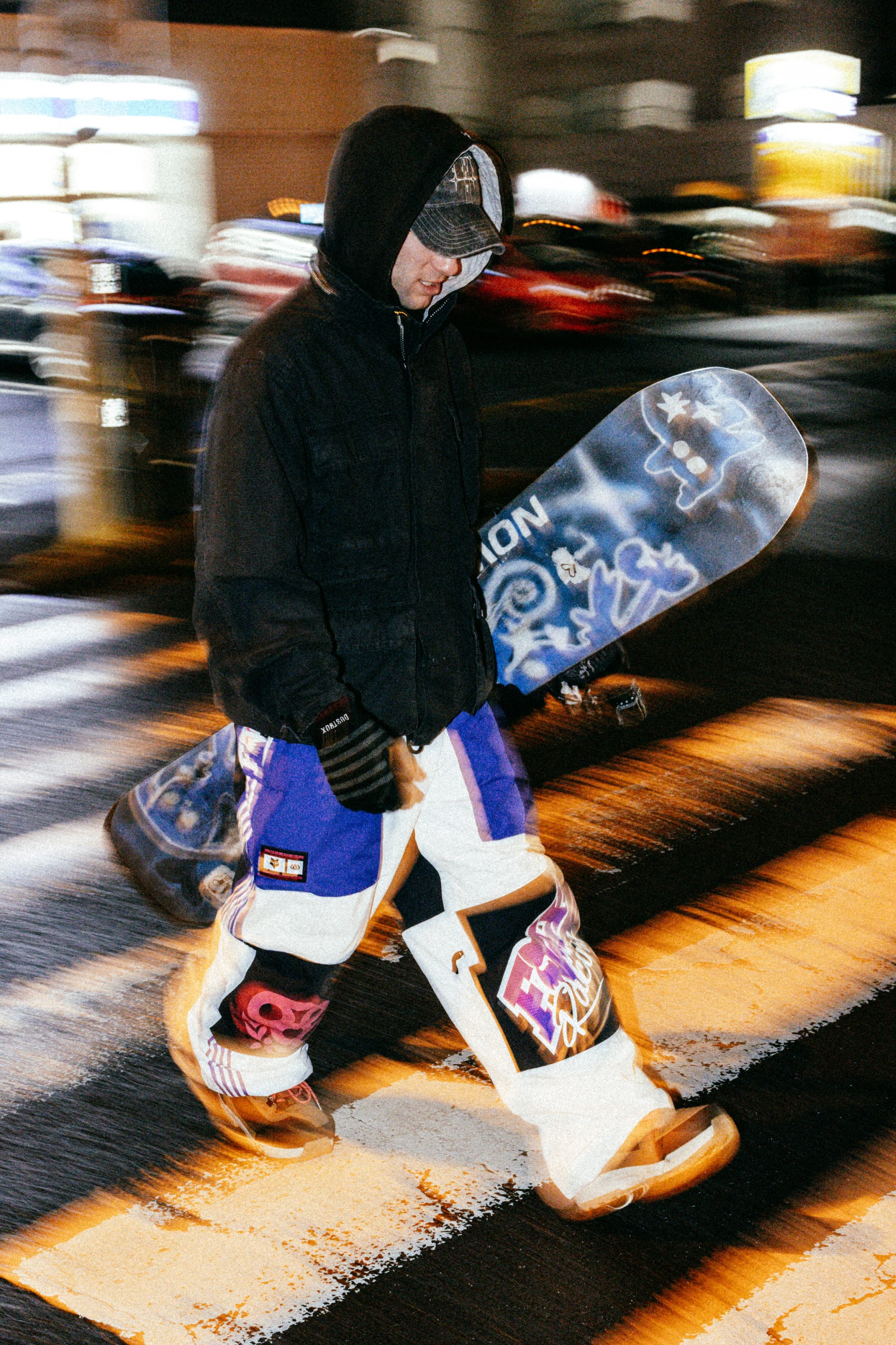 Wearing a hooded jacket and 686 Men's Fox Racing Shell Pant, a person walks across a city street at night, carrying a snowboard with blue and white graphics. Blurred city lights fill the background.