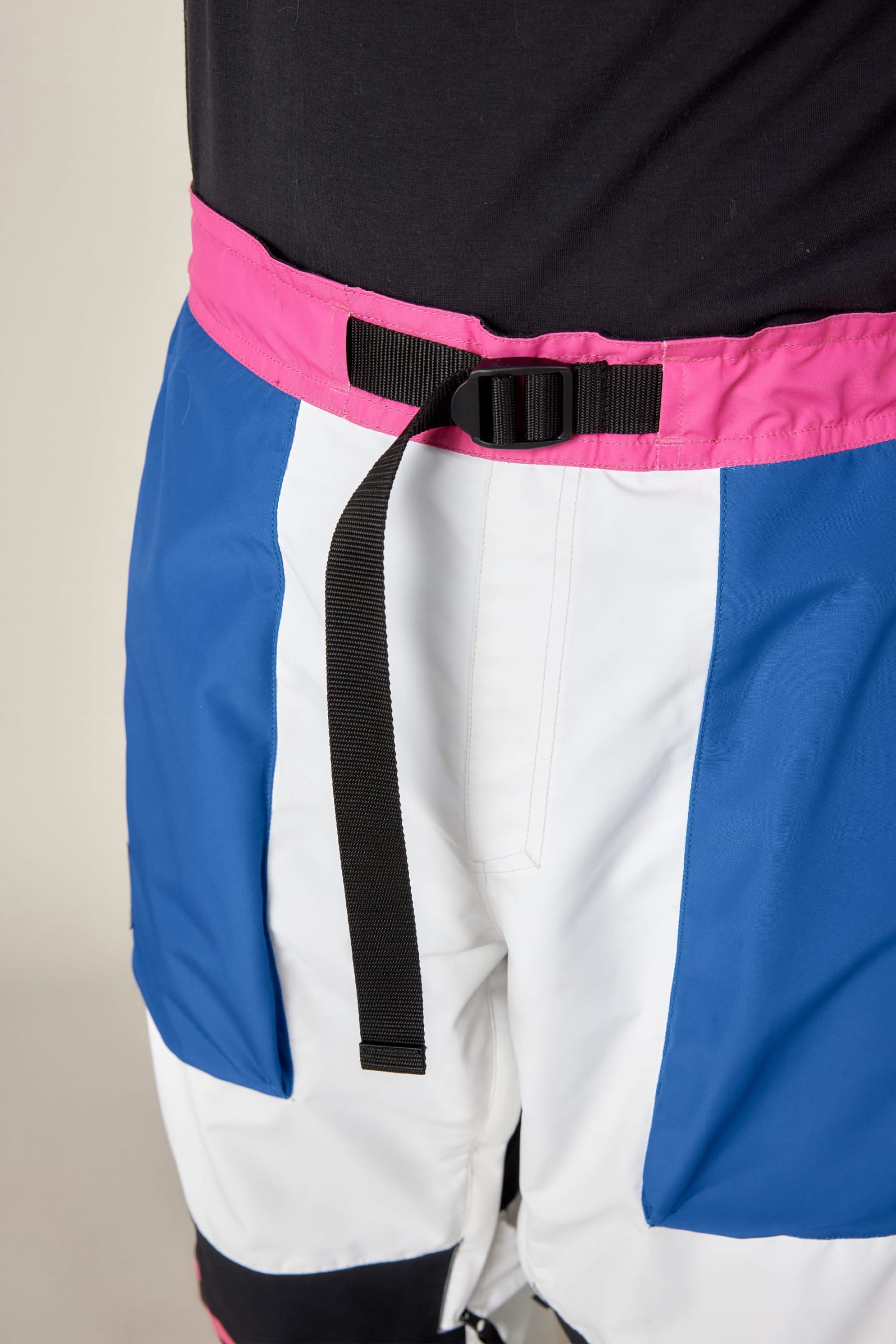 A close-up of a person wearing the 686 Men's Fox Racing Shell Pant by 686, featuring color-blocked blue and white panels, a pink waistband, a black adjustable belt, and paired with a black top.