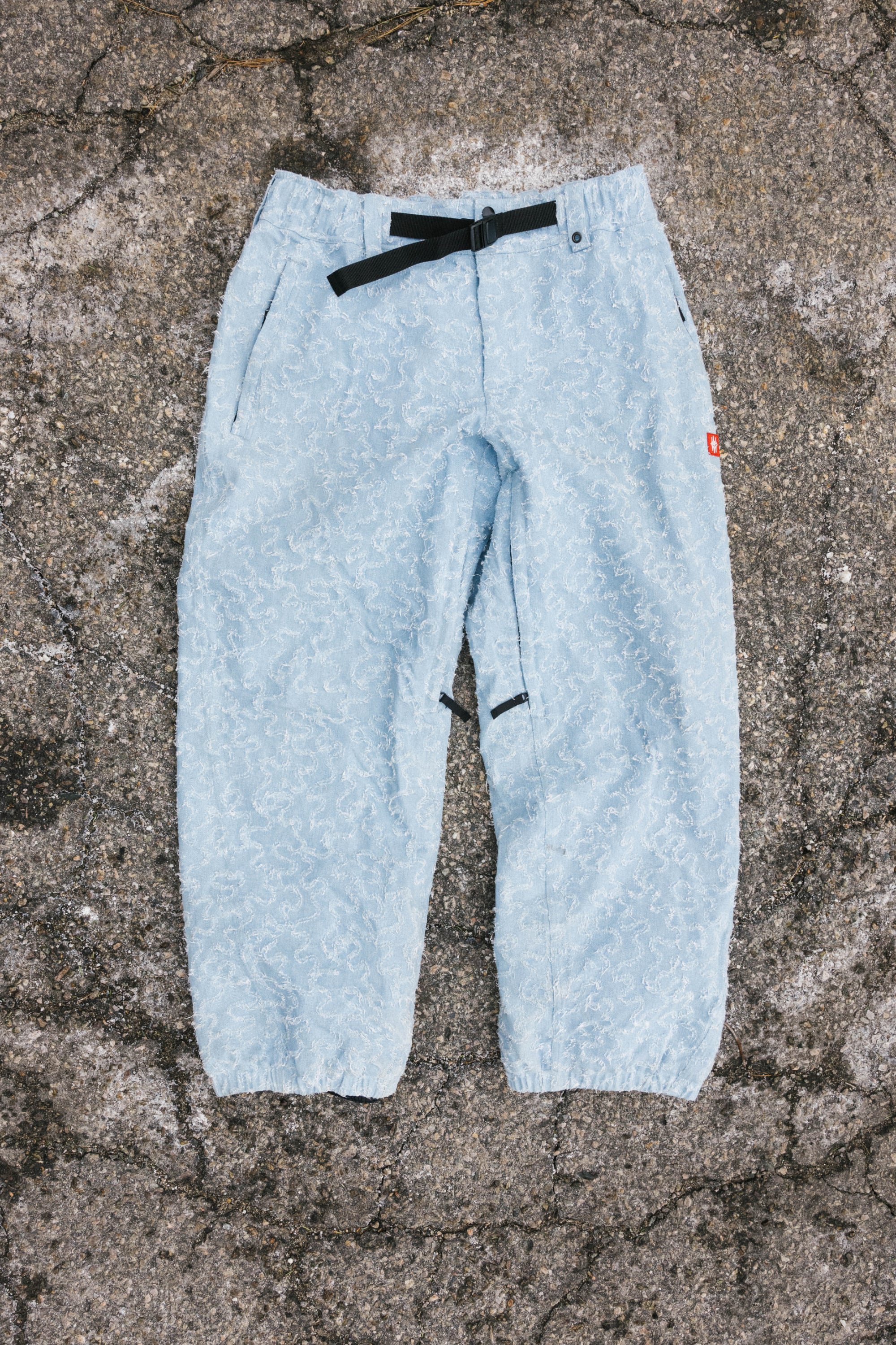 The 686 Dojo® Frayed Denim Pant by 686, shown in light blue with an adjustable black belt and elastic cuffs, is displayed flat on a rough, cracked concrete surface.