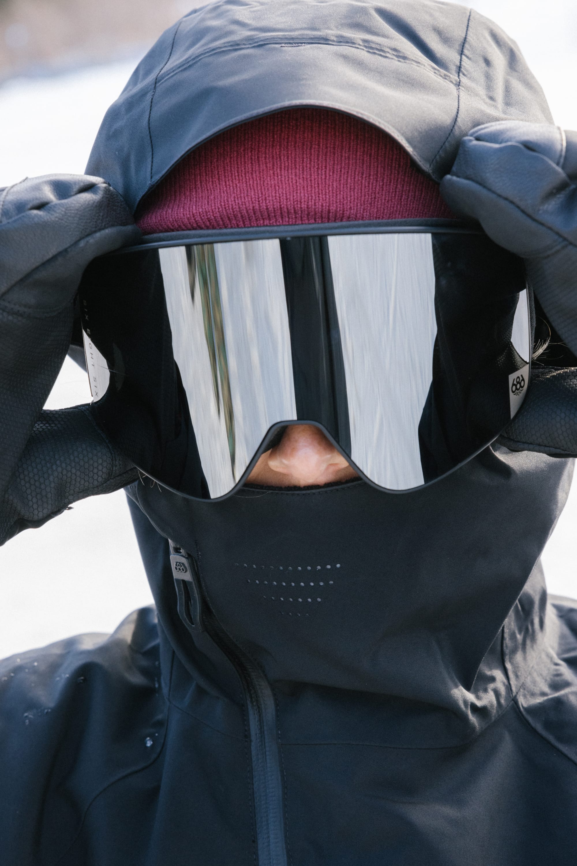 A person in the 686 Women's Hydra™ Thermagraph® Jacket adjusts their ski goggles, wearing a maroon beanie and black gloves while standing in the snow.