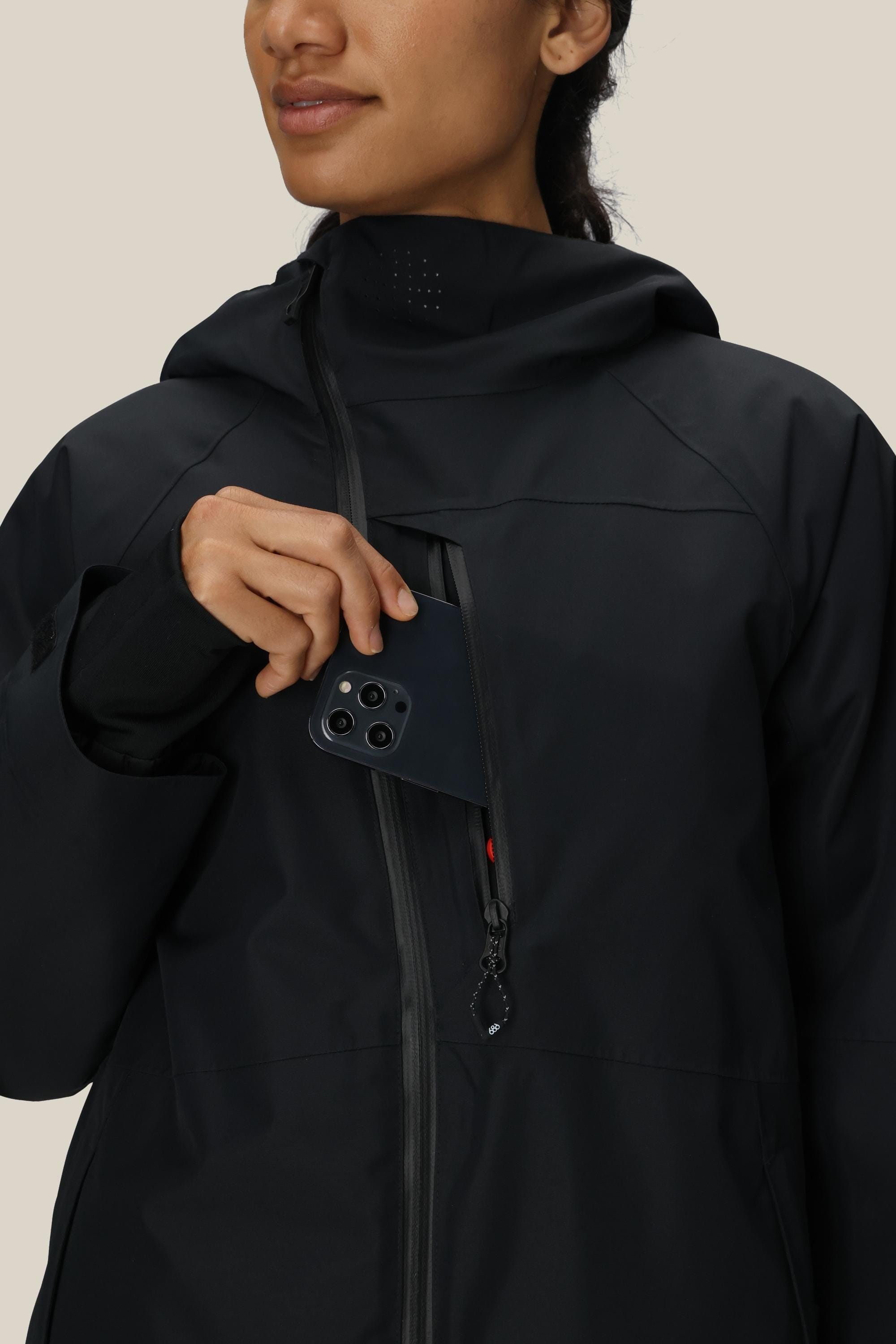 A woman in a 686 Women's Hydra™ Thermagraph® Jacket by 686 unzips the upper chest pocket to place a black smartphone inside. Her face is partially visible against a plain, light-colored background.