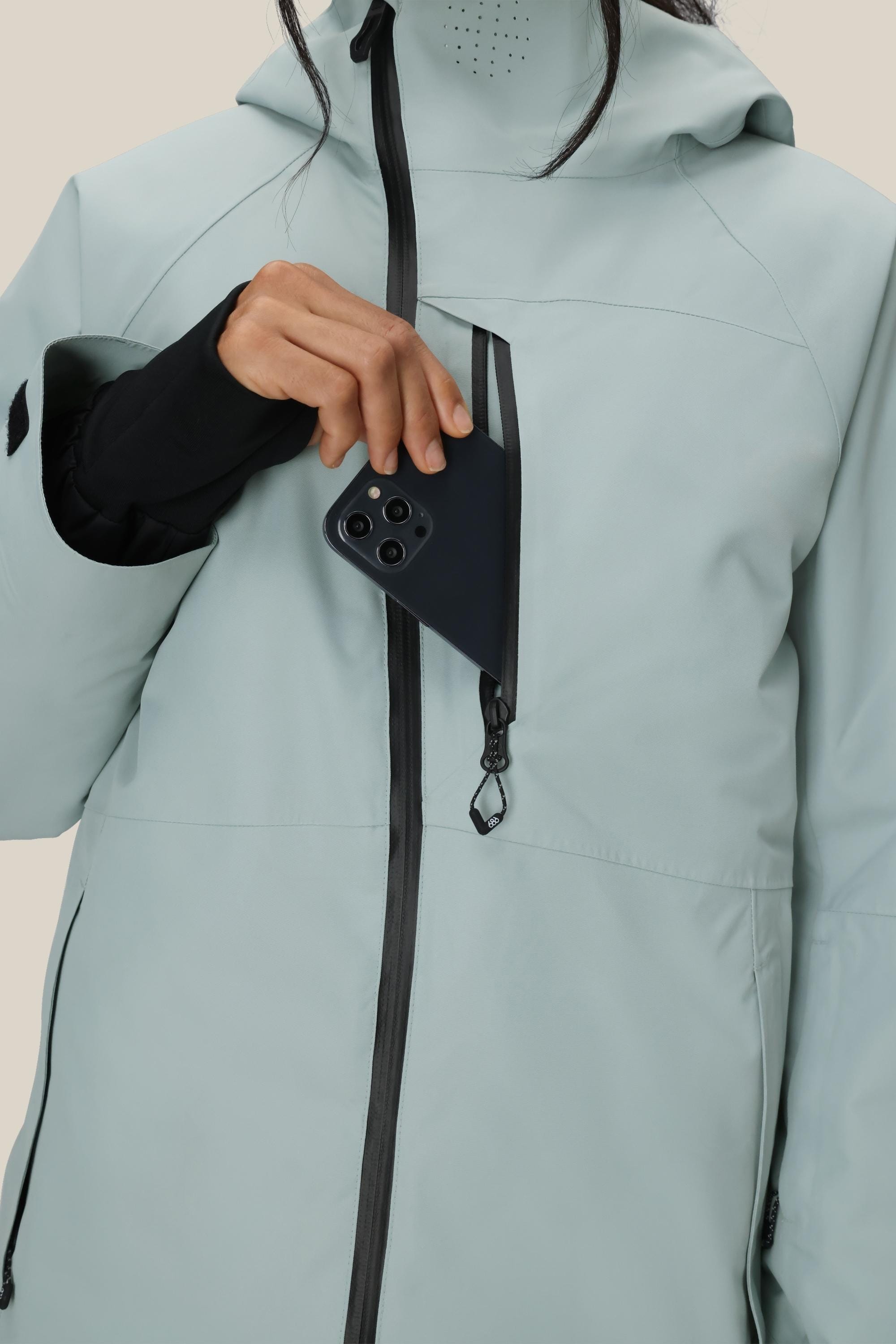 A woman wearing the 686 Women's Hydra™ Thermagraph® Jacket in light gray slips a black smartphone into the jacket's zippered chest pocket.