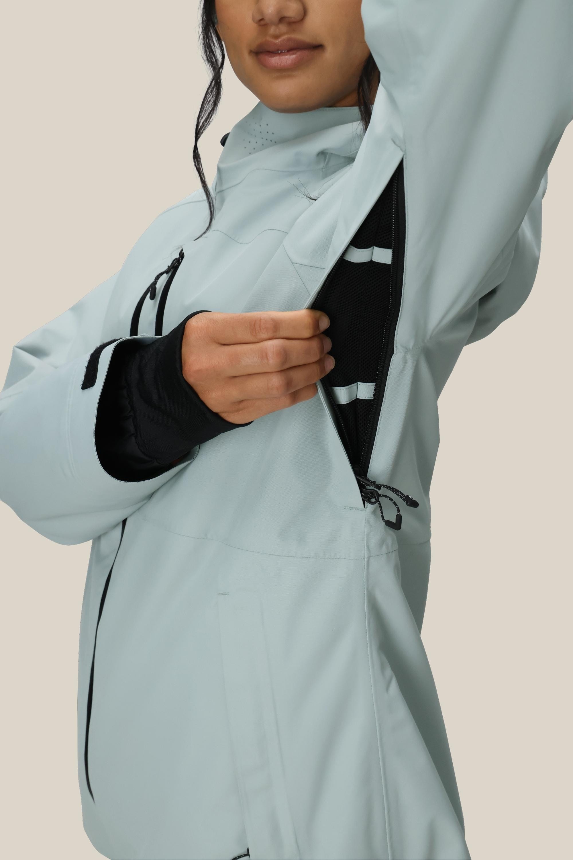 A woman in the 686 Women's Hydra™ Thermagraph® Jacket by 686 unzips an underarm vent to reveal black mesh lining. Her face is partly visible against a plain beige background.