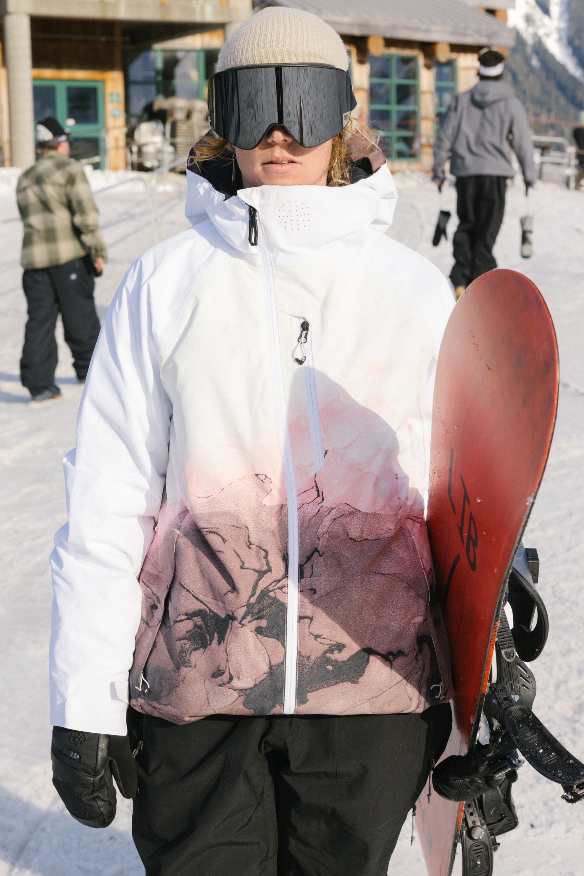 Wearing a 686 Women's Hydra™ Thermagraph® Jacket with white and pink patterns, black pants, gloves, helmet, and reflective ski goggles, a person holds a red snowboard on a snowy slope with others and a lodge in the background.