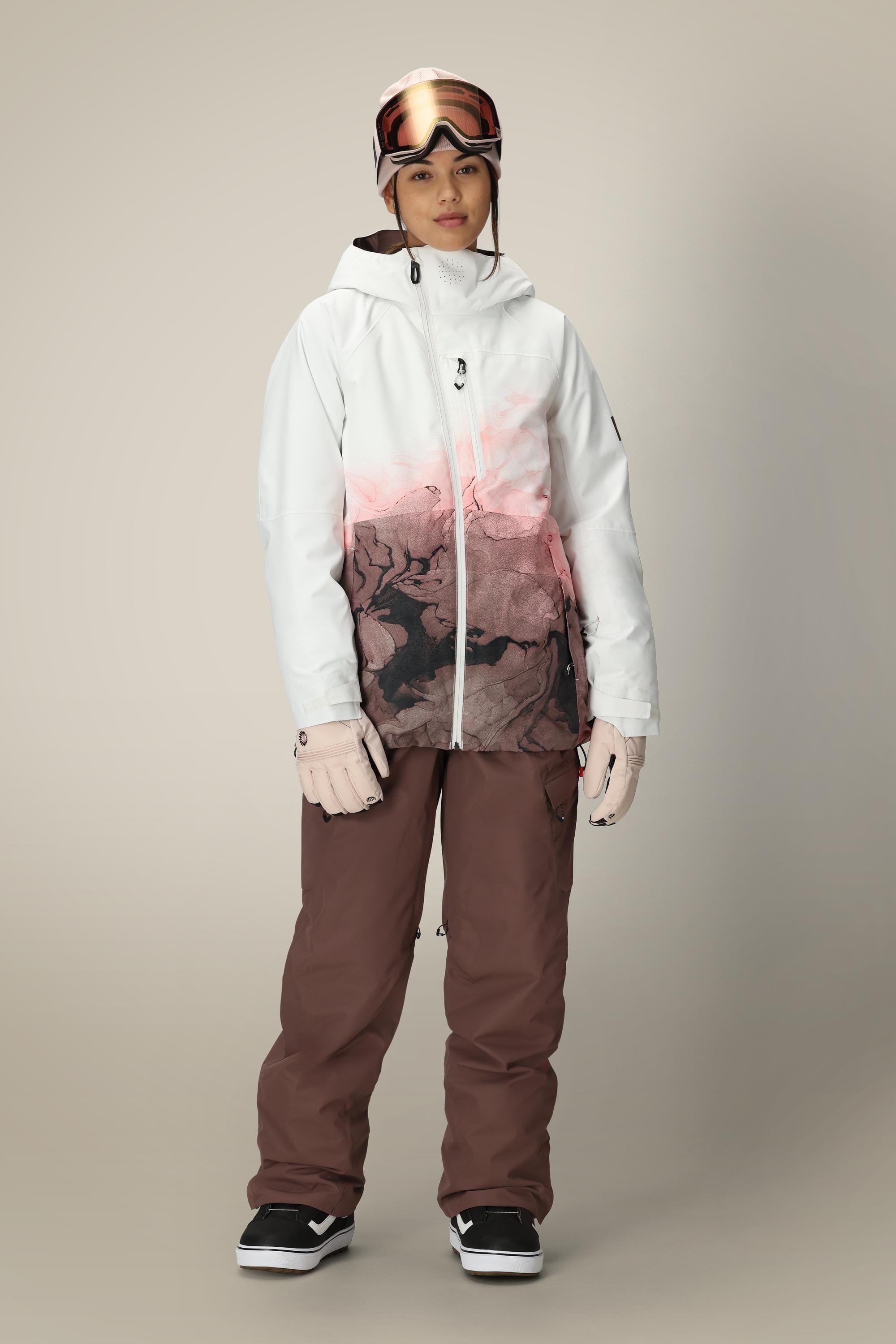 A person stands facing forward against a plain background, wearing the 686 Women's Hydra™ Thermagraph® Jacket with a black abstract design in white and pink, brown snow pants, white gloves, black sneakers, and ski goggles.