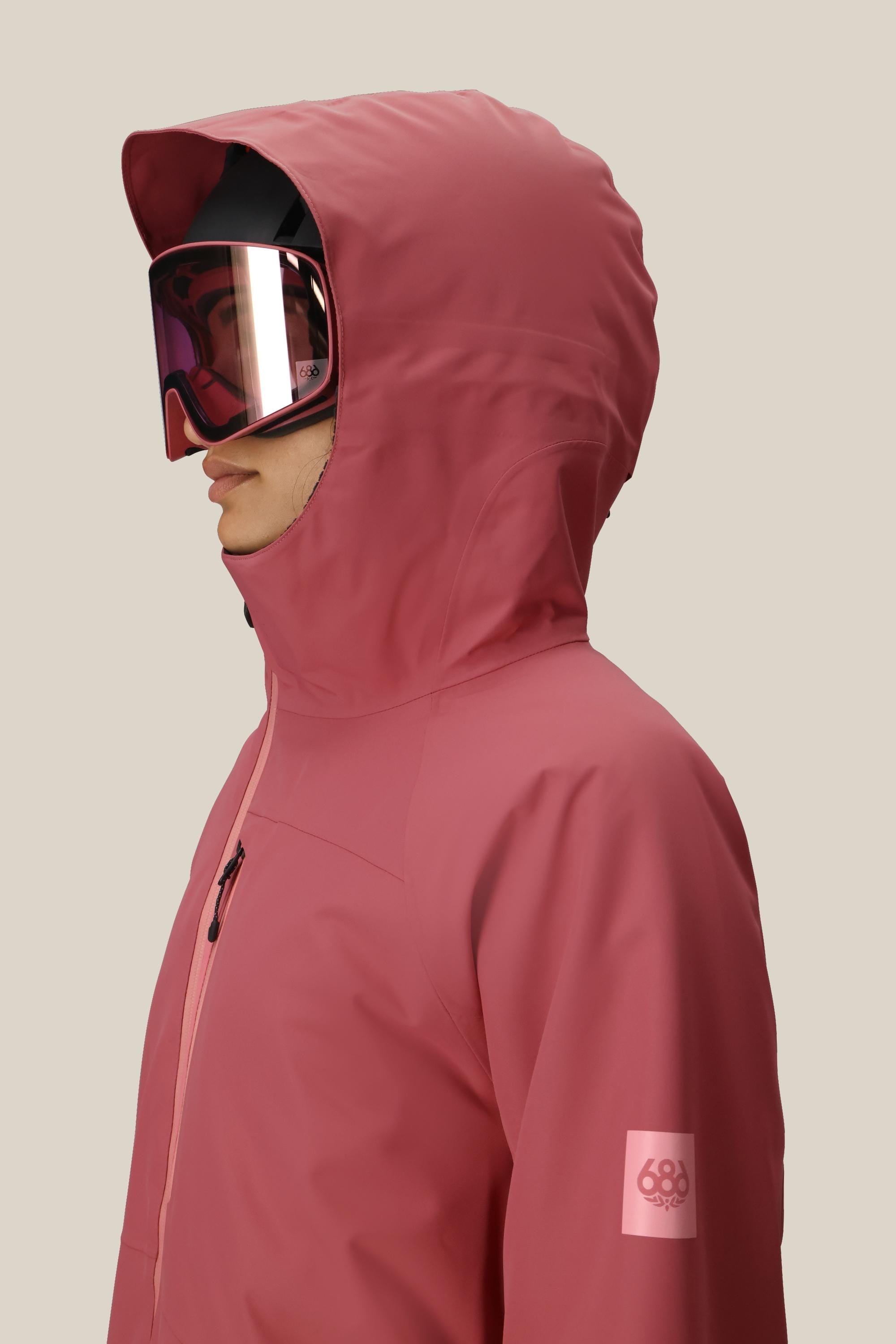 A person in a pink 686 Women's Hydra™ Thermagraph® Jacket and pink ski goggles faces left against a beige background. The waterproof jacket, by 686, displays the brand's logo on the sleeve.
