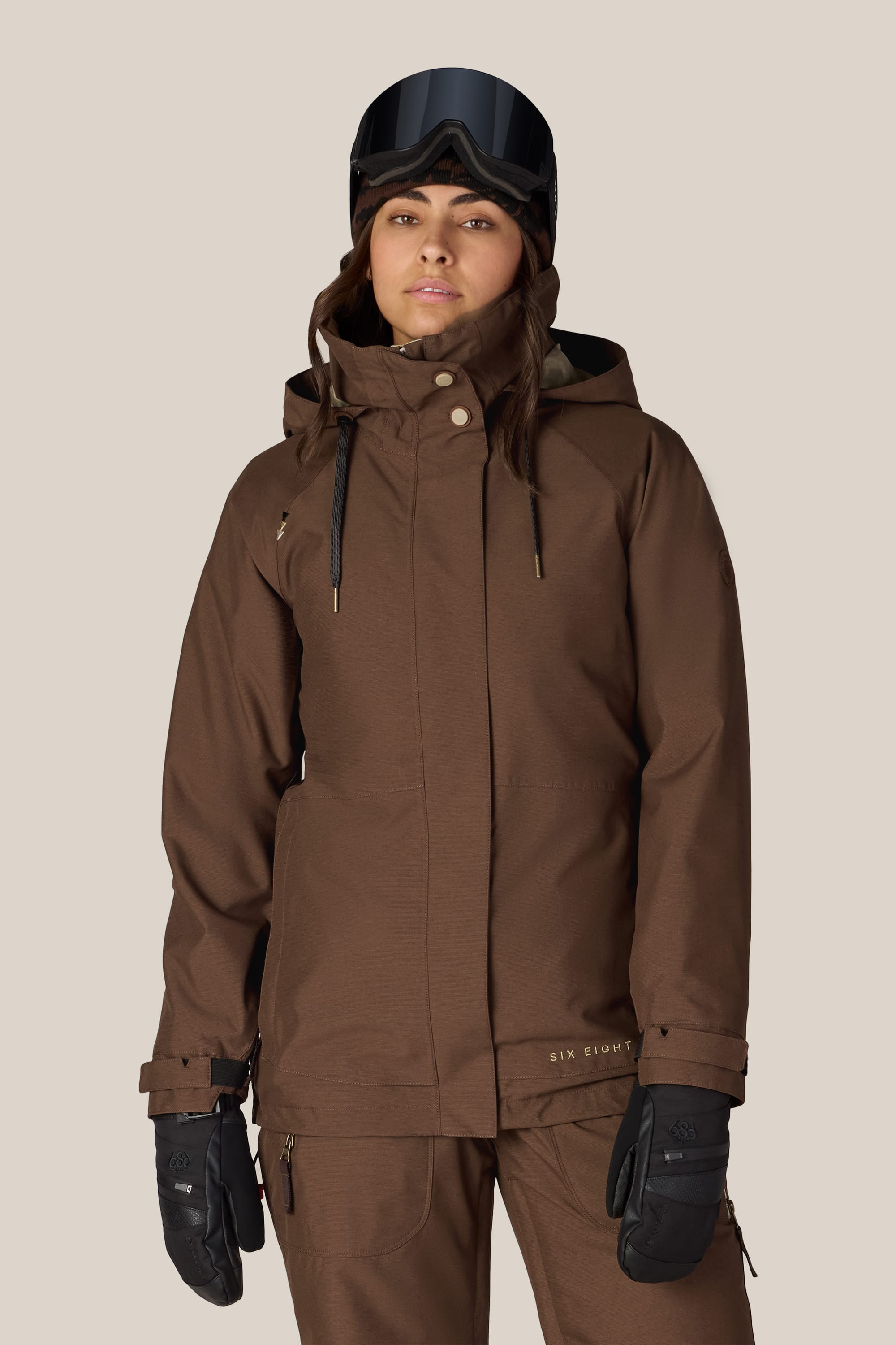 A woman dressed in the 686 Women’s SMARTY® 3-In-1 Spellbound Jacket stands against a plain light background, paired with matching pants, black gloves, and black ski goggles.