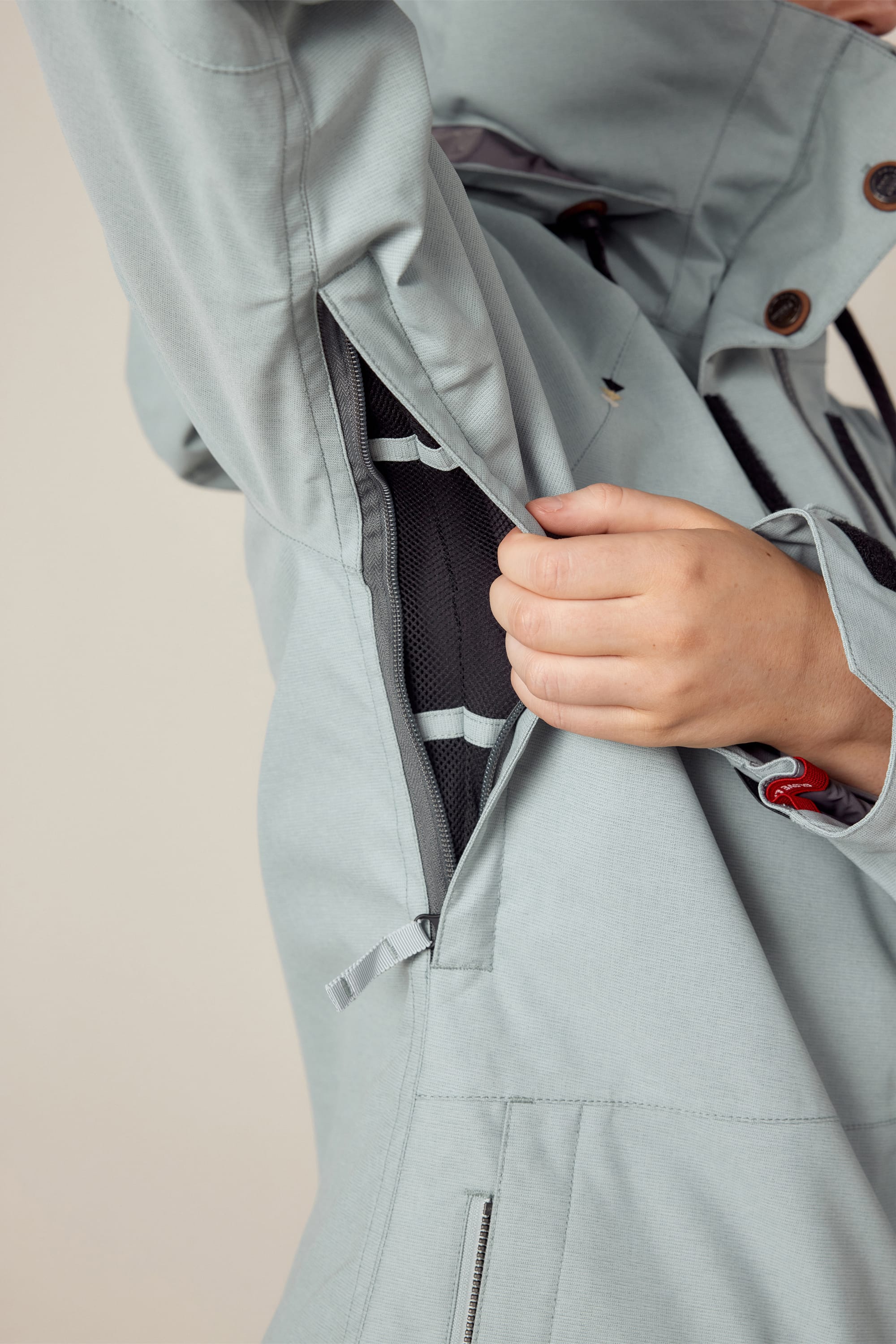 A person unzips the underarm vent of the 686 Women's SMARTY® 3-In-1 Spellbound Jacket, revealing a black mesh lining inside the light gray polyester shell.