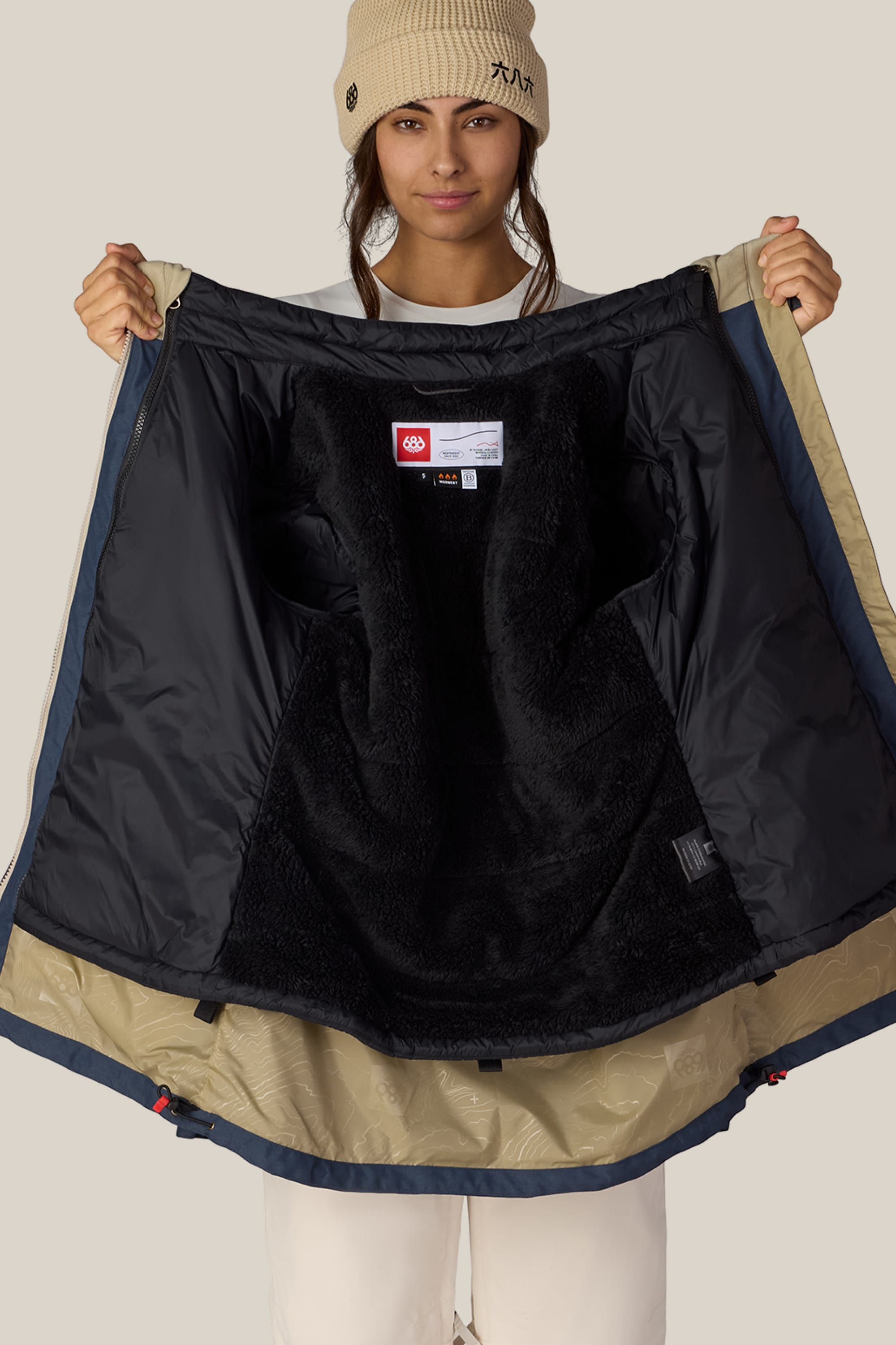 A person wearing a tan beanie opens the 686 Women’s SMARTY® 3-In-1 Spellbound Jacket to reveal its soft, furry black lining and interior labels, standing against a plain background.