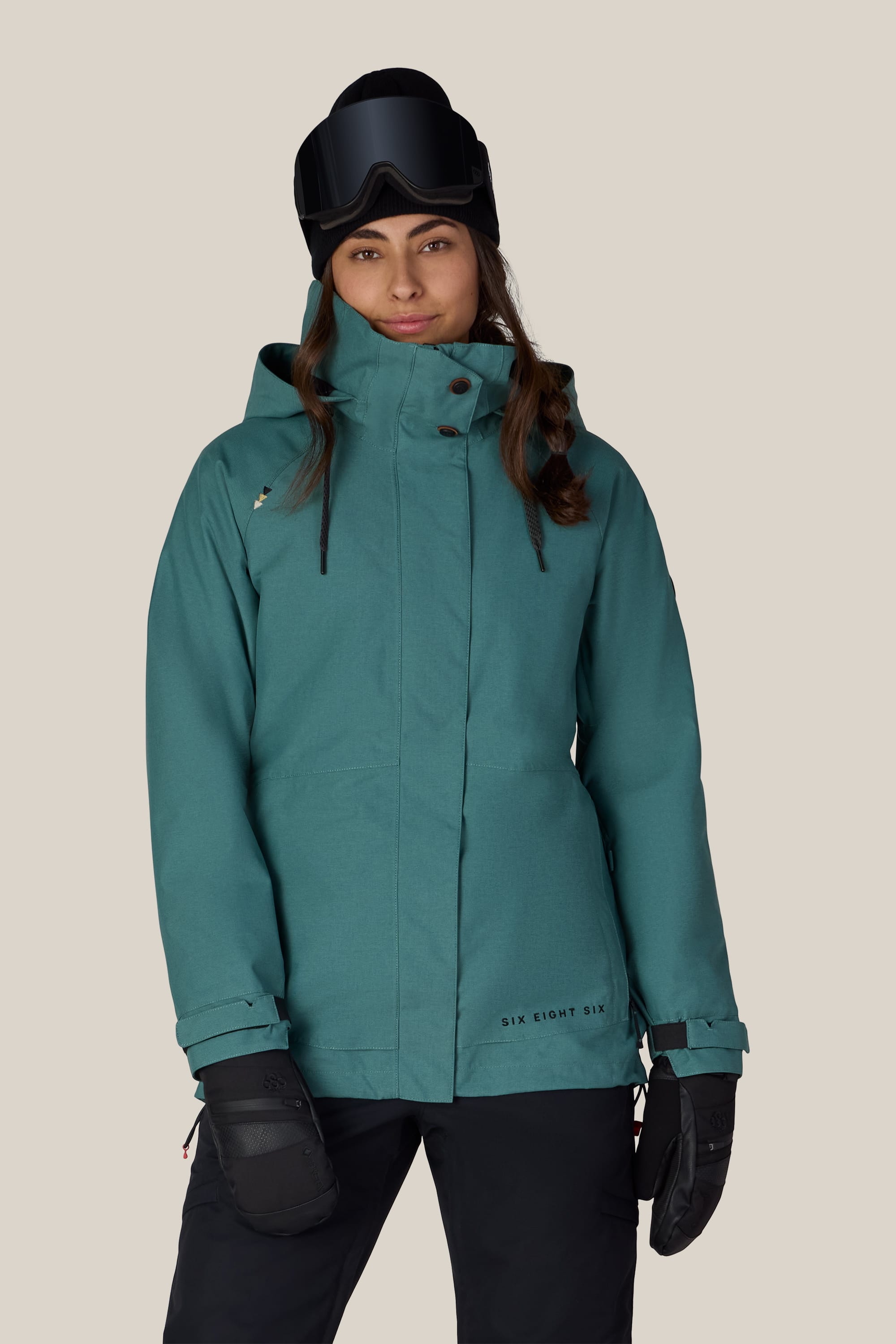A person wearing the 686 Women’s SMARTY® 3-In-1 Spellbound Jacket in teal, paired with black snow pants, black gloves, and ski goggles stands facing forward against a plain light background.