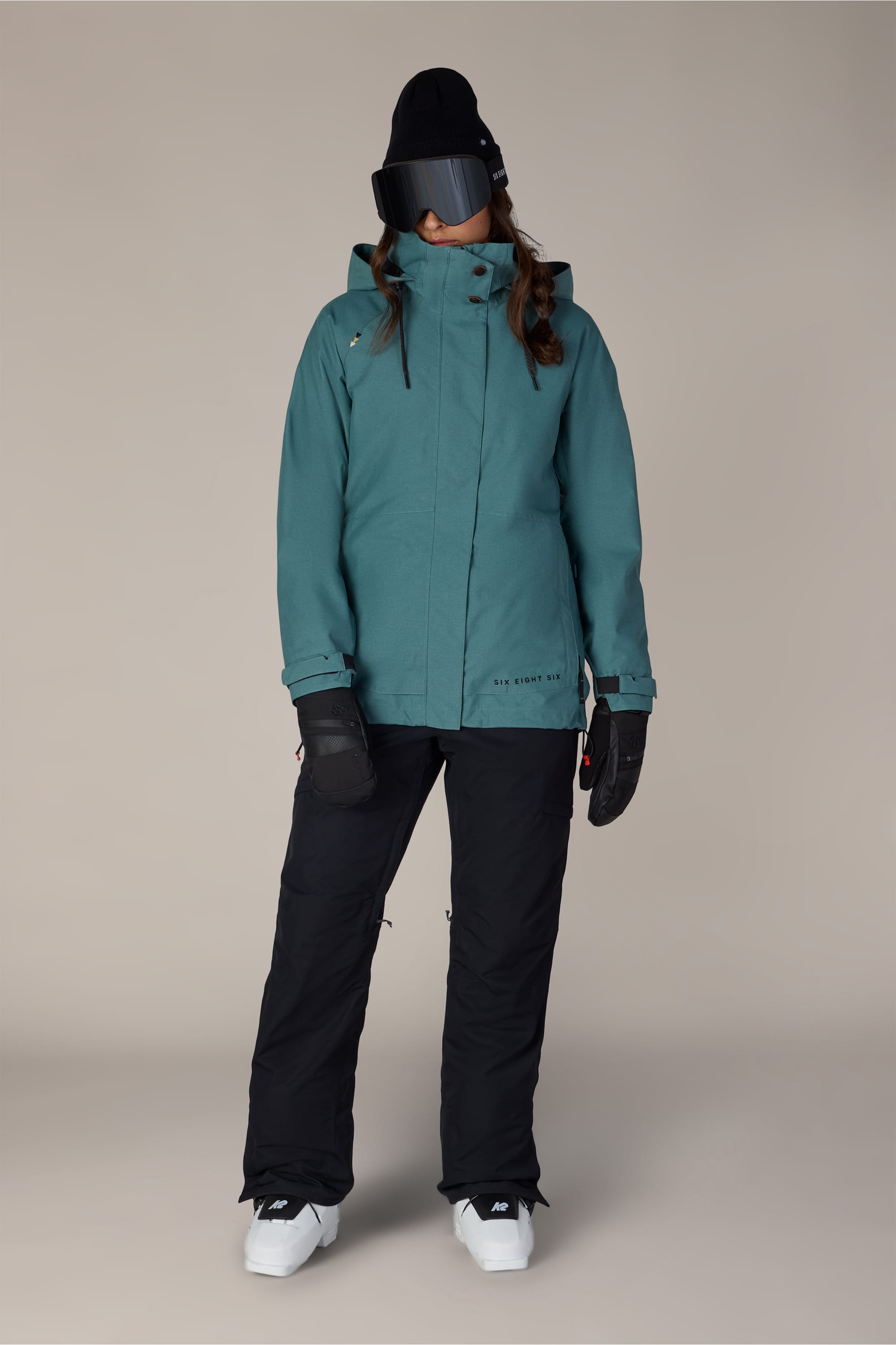 A person wearing the 686 Women’s SMARTY® 3-In-1 Spellbound Jacket by 686, along with black snow pants, black gloves, a black beanie, dark ski goggles, and white boots stands against a plain background.