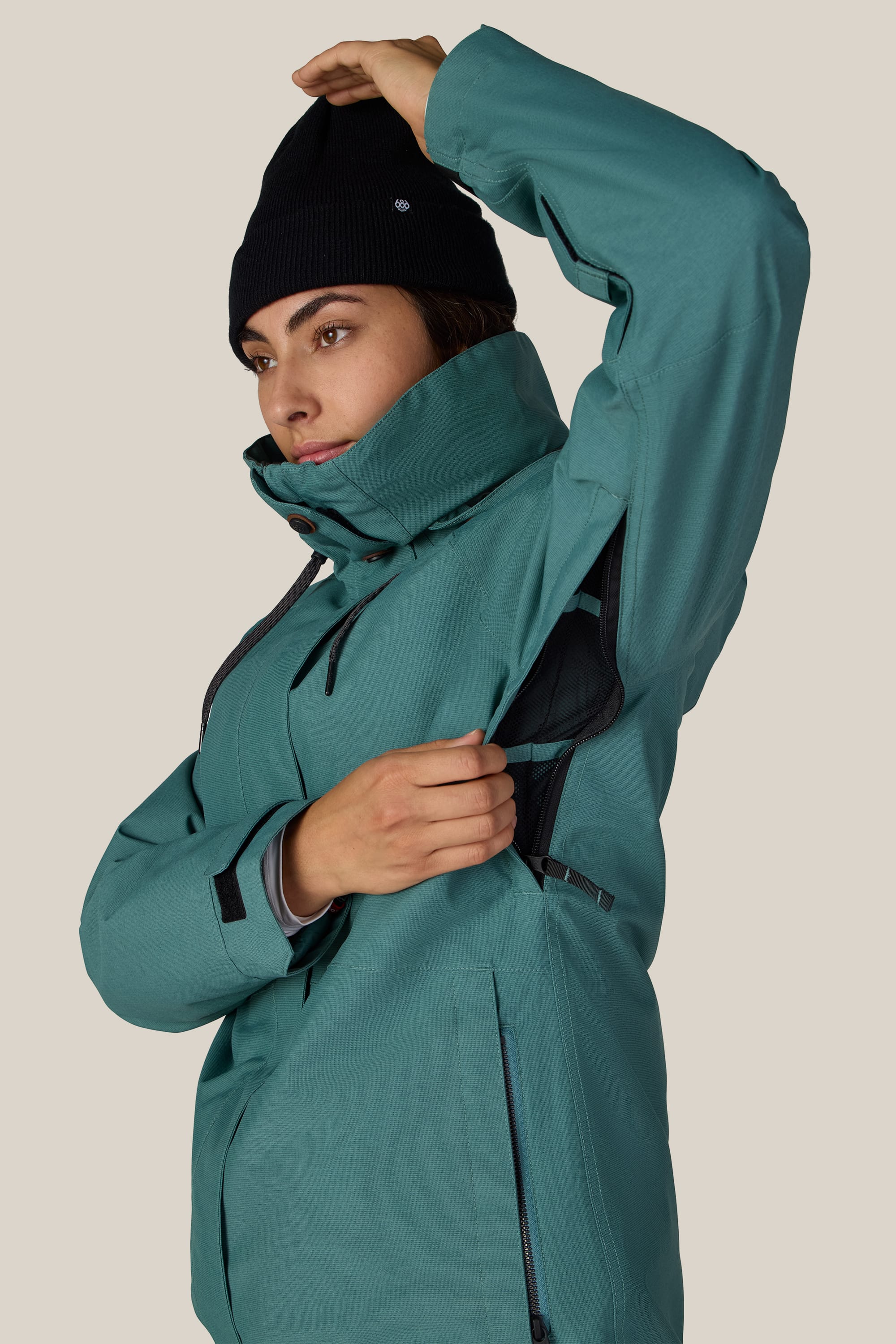 A person wearing the 686 Women’s SMARTY® 3-In-1 Spellbound Jacket in teal with a black beanie lifts their arm, revealing a zippered sleeve vent against a plain background.