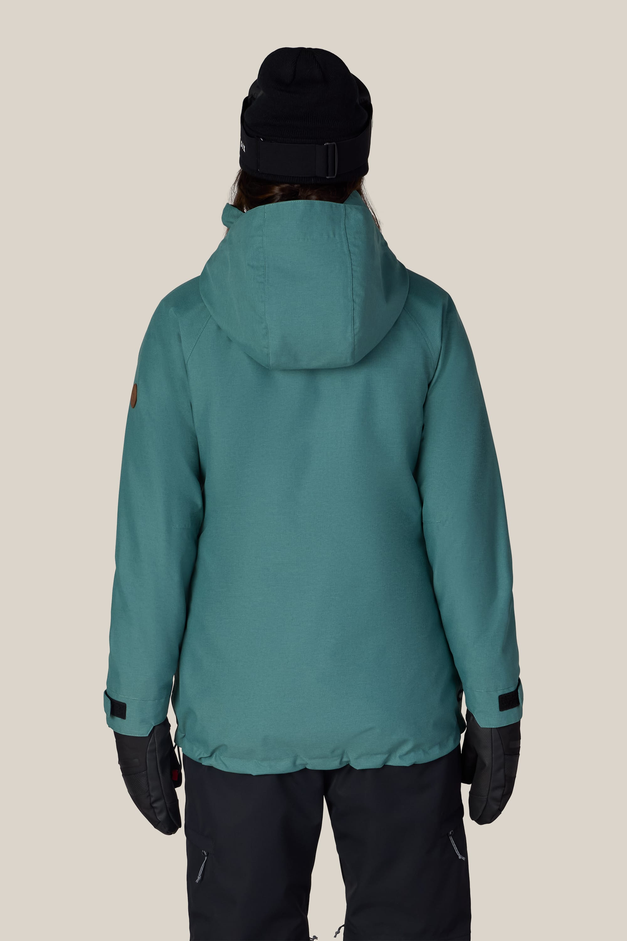 A person is seen from behind, wearing the 686 Women’s SMARTY® 3-In-1 Spellbound Jacket in teal, paired with a black beanie, black gloves, and black pants, set against a plain beige background.