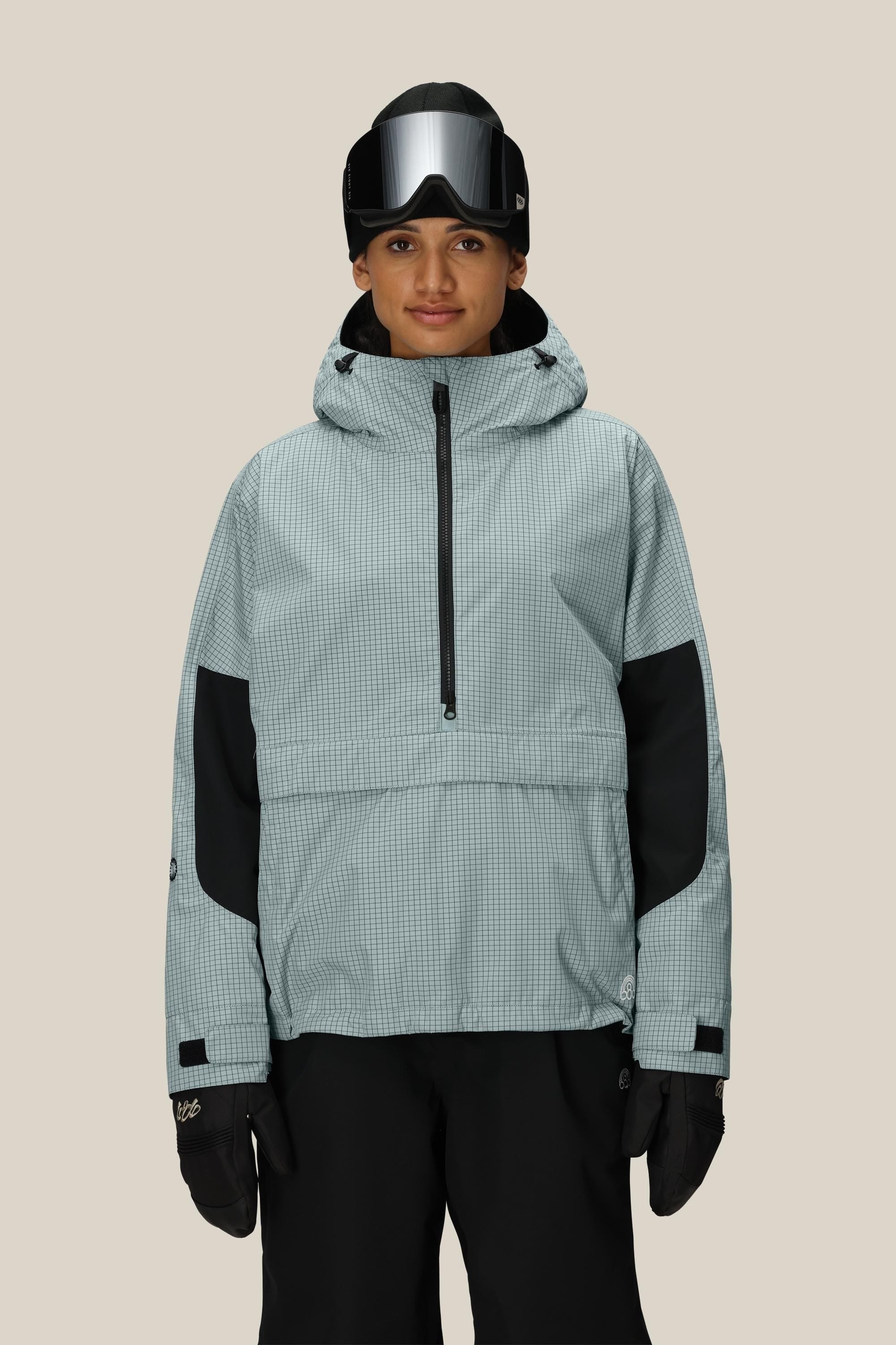 A person wearing the 686 Women's Outline™ Shell Anorak in light gray with black accents, black ski gloves, black pants, and ski goggles on their forehead stands against a plain beige background.