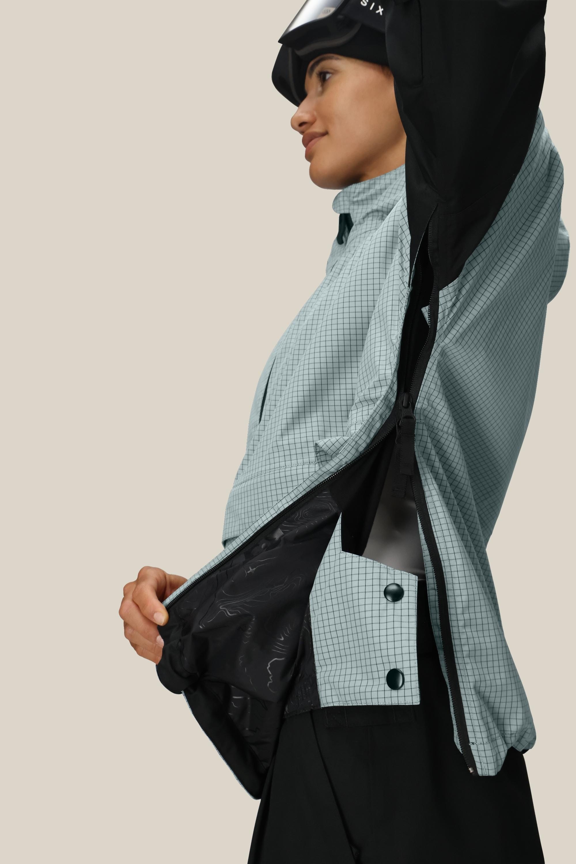 A woman in a light blue checkered jacket unzips the 686 Women's Outline™ Shell Anorak, revealing its inner lining. She is viewed from the side with a slight smile, all set against a plain beige background.