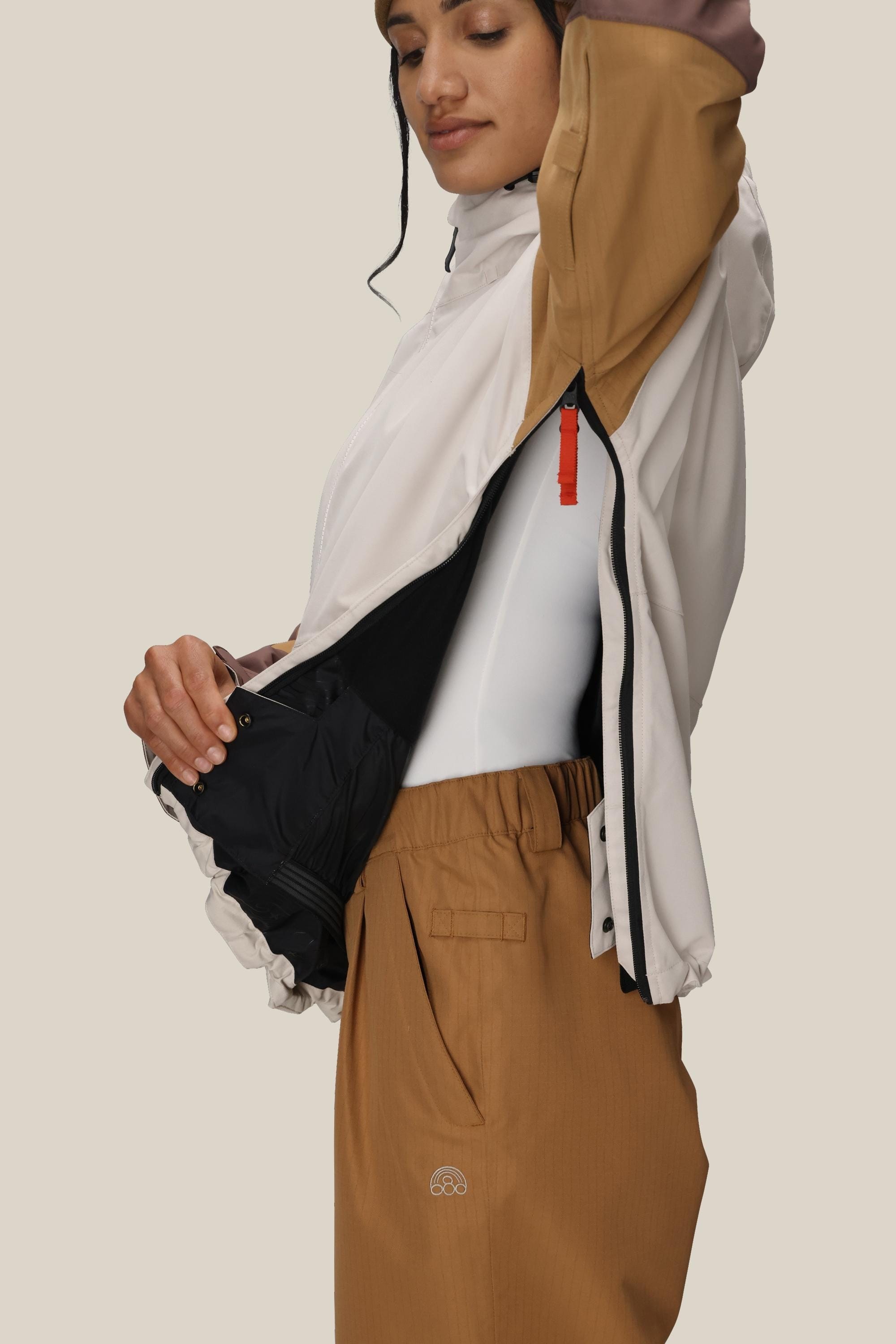 A person in brown pants and a 686 Women's Outline™ Shell Anorak with black lining unzips the side, revealing a white shirt underneath. The background is plain beige.