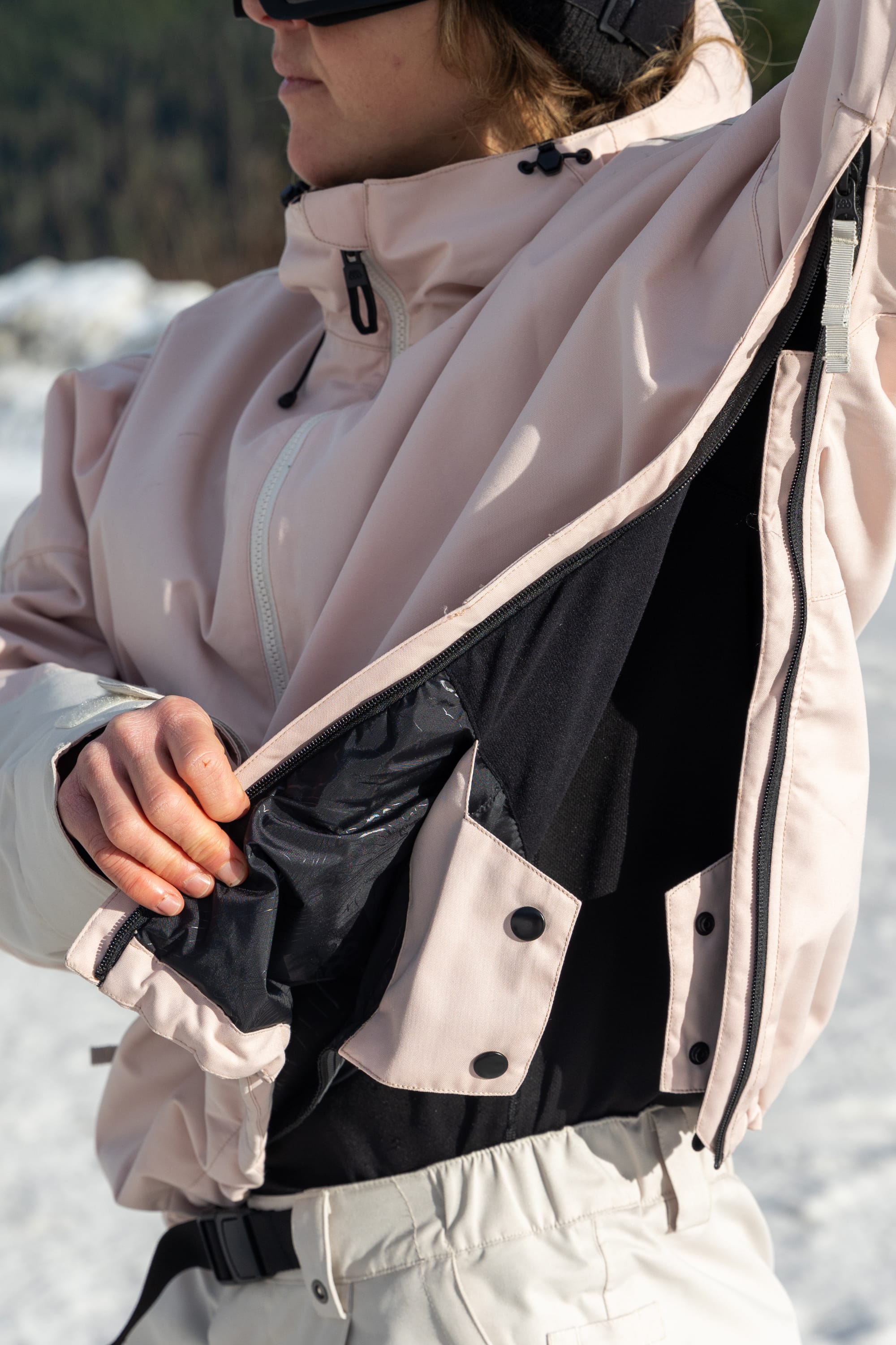 A woman in a 686 Women's Outline™ Shell Anorak in light pink with white pants opens a zippered side vent to reveal black lining while standing outdoors in the snow.