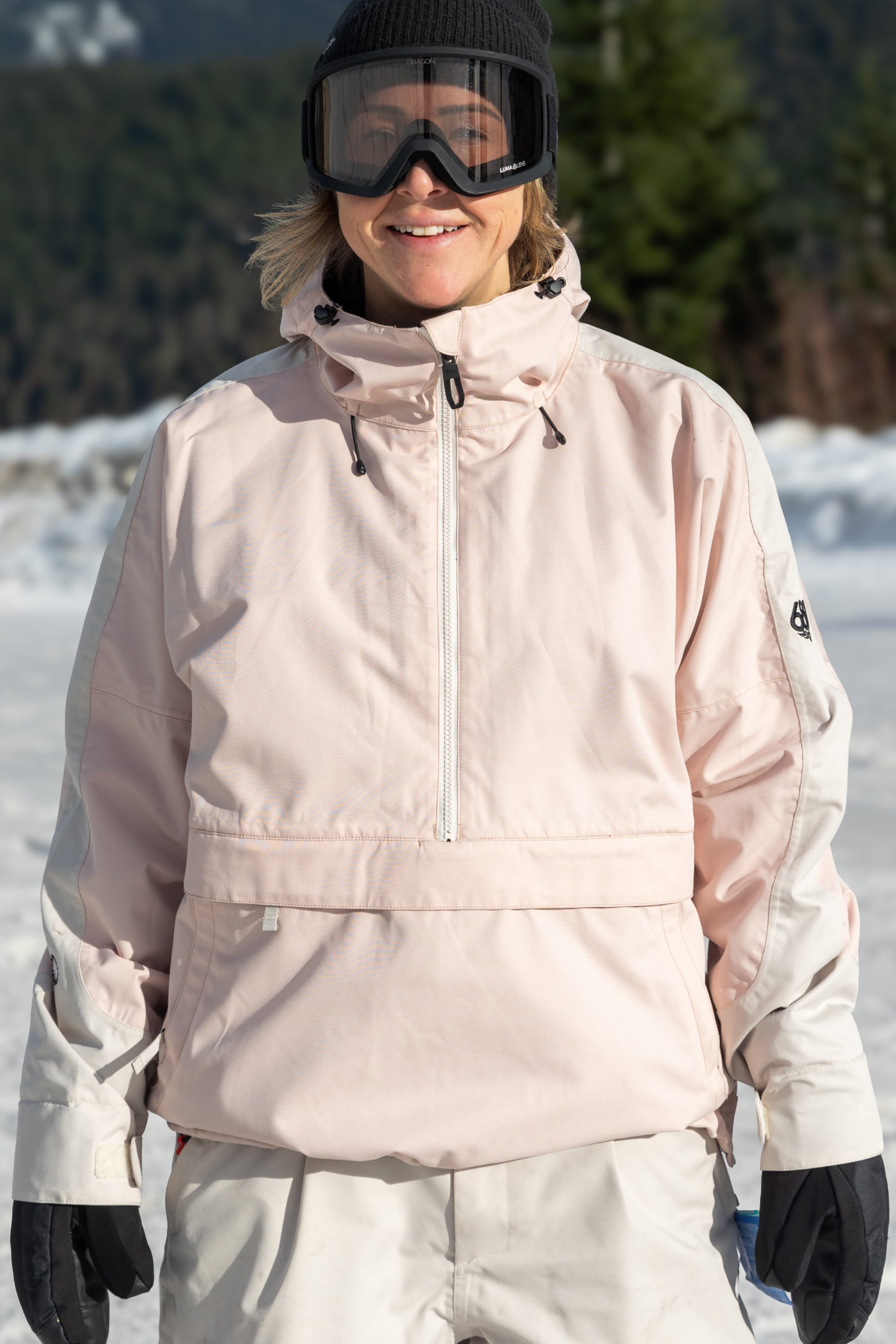 Wearing a 686 Women's Outline™ Shell Anorak, white pants, black gloves, and black ski goggles, a person smiles outdoors in a snowy landscape with trees in the background.