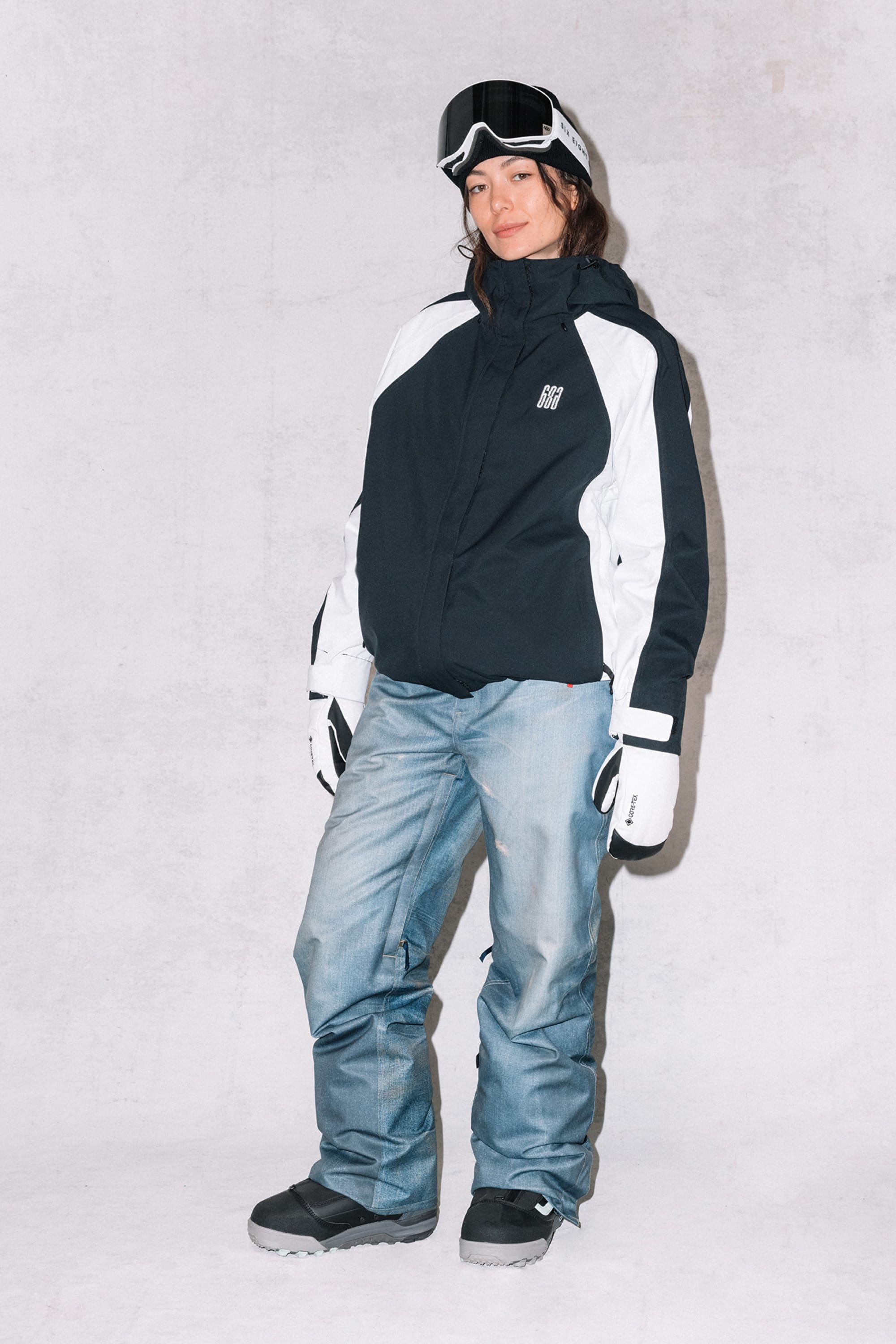 A person wearing the 686 Women's Hologram™ Shell Jacket, blue snow pants, black snow boots, white gloves, and ski goggles poses against a light background for the camera.
