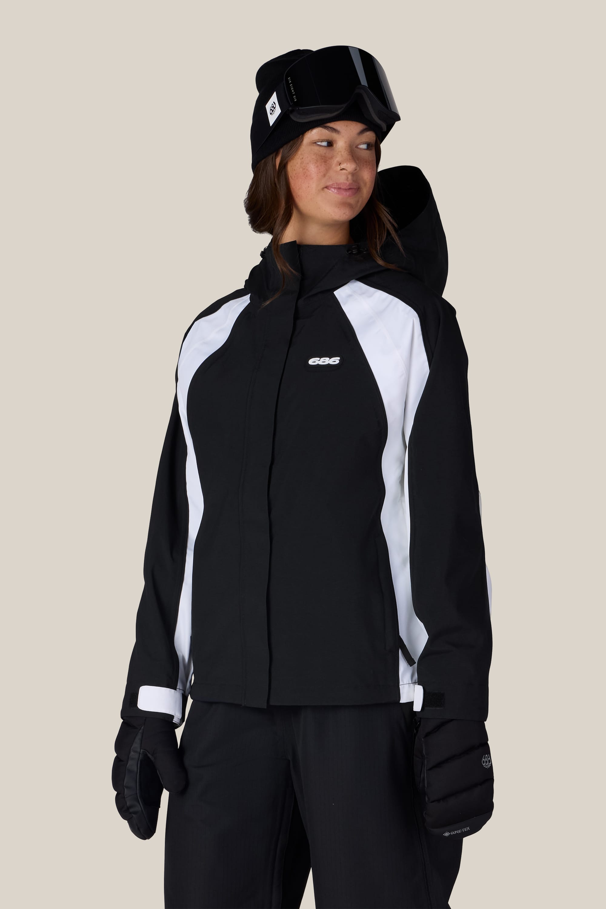 Wearing the 686 Women’s Hologram™ Shell Jacket from 686 in black and white, along with black pants, gloves, a beanie, and ski goggles, a person stands against a plain light background, looking to the side with a slight smile.