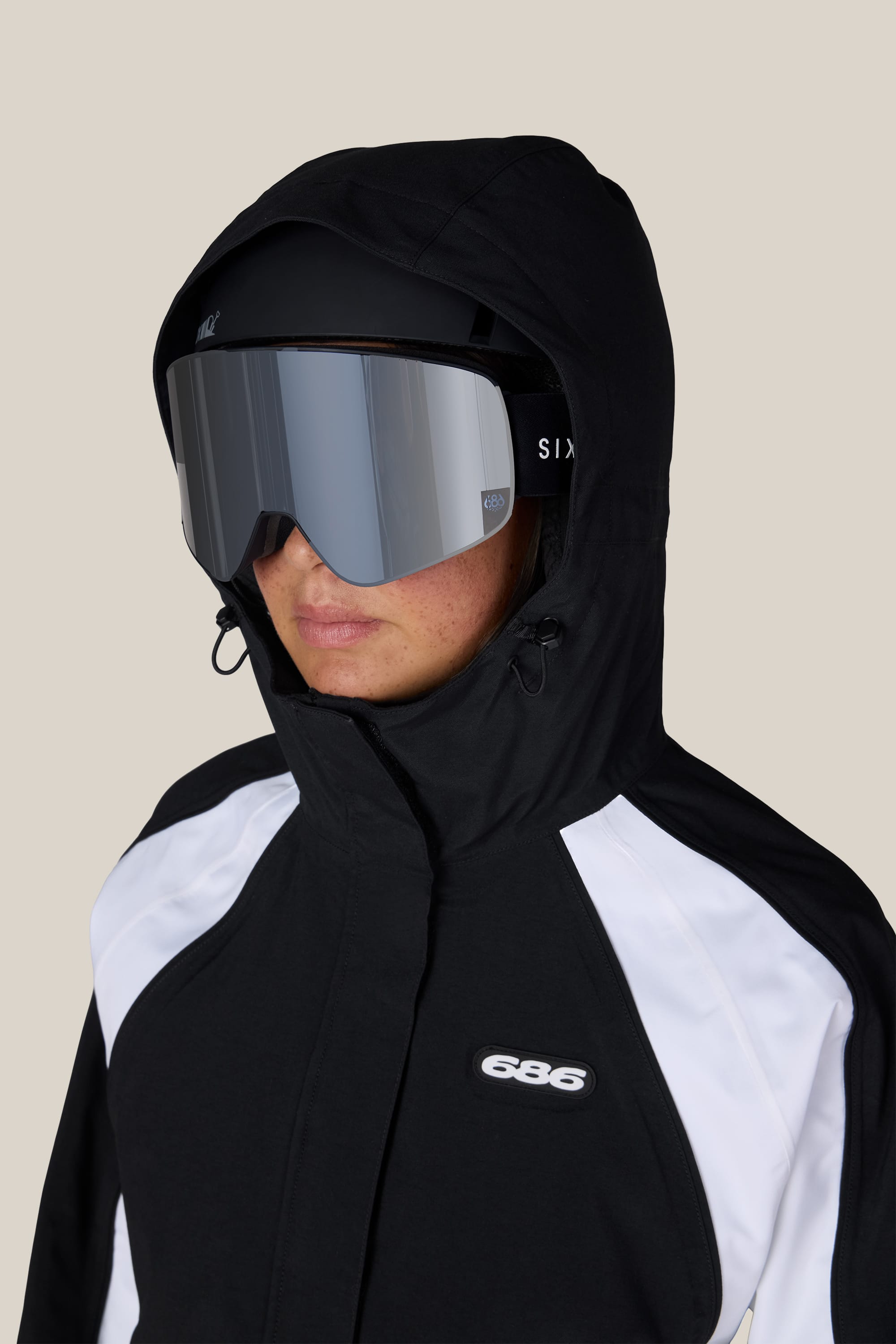 A person wearing the 686 Women’s Hologram™ Shell Jacket by 686, paired with large reflective ski goggles and a black helmet, stands against a plain beige background.