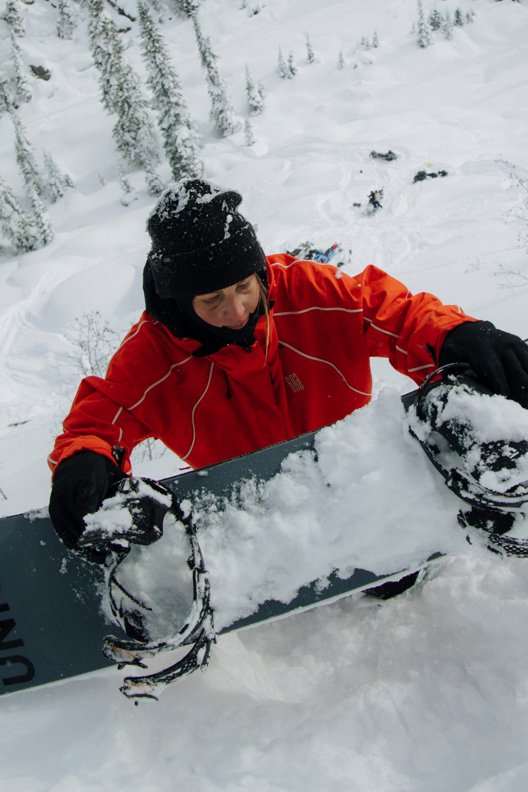 Wearing the 686 Women's Hologram™ Shell Jacket, a snowboarder in a black hat climbs a snowy hill, carrying a snow-covered snowboard amid wintry trees and landscape.