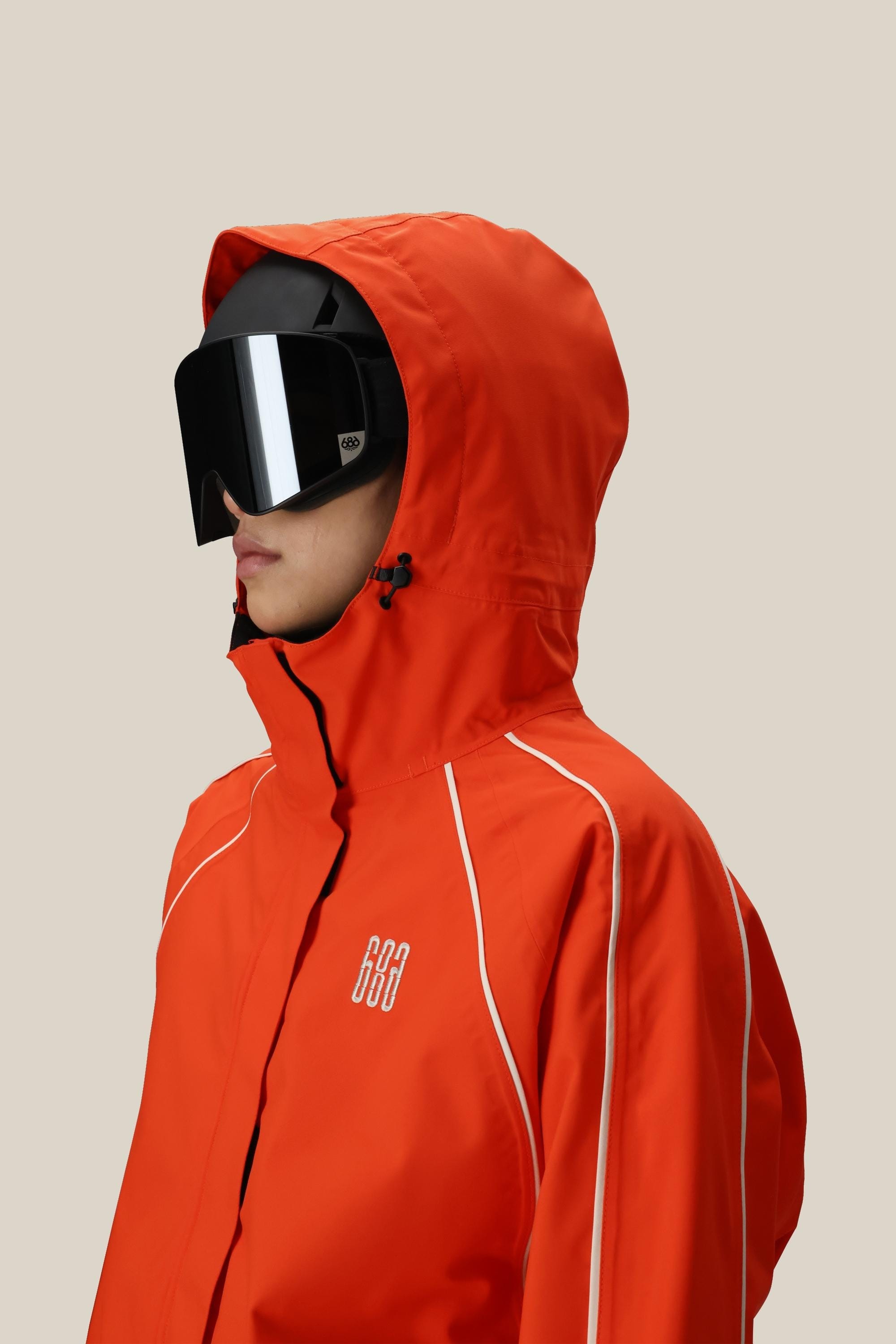 A person wears the 686 Women's Hologram™ Shell Jacket in bright orange with white piping and large black ski goggles, standing against a plain beige background.