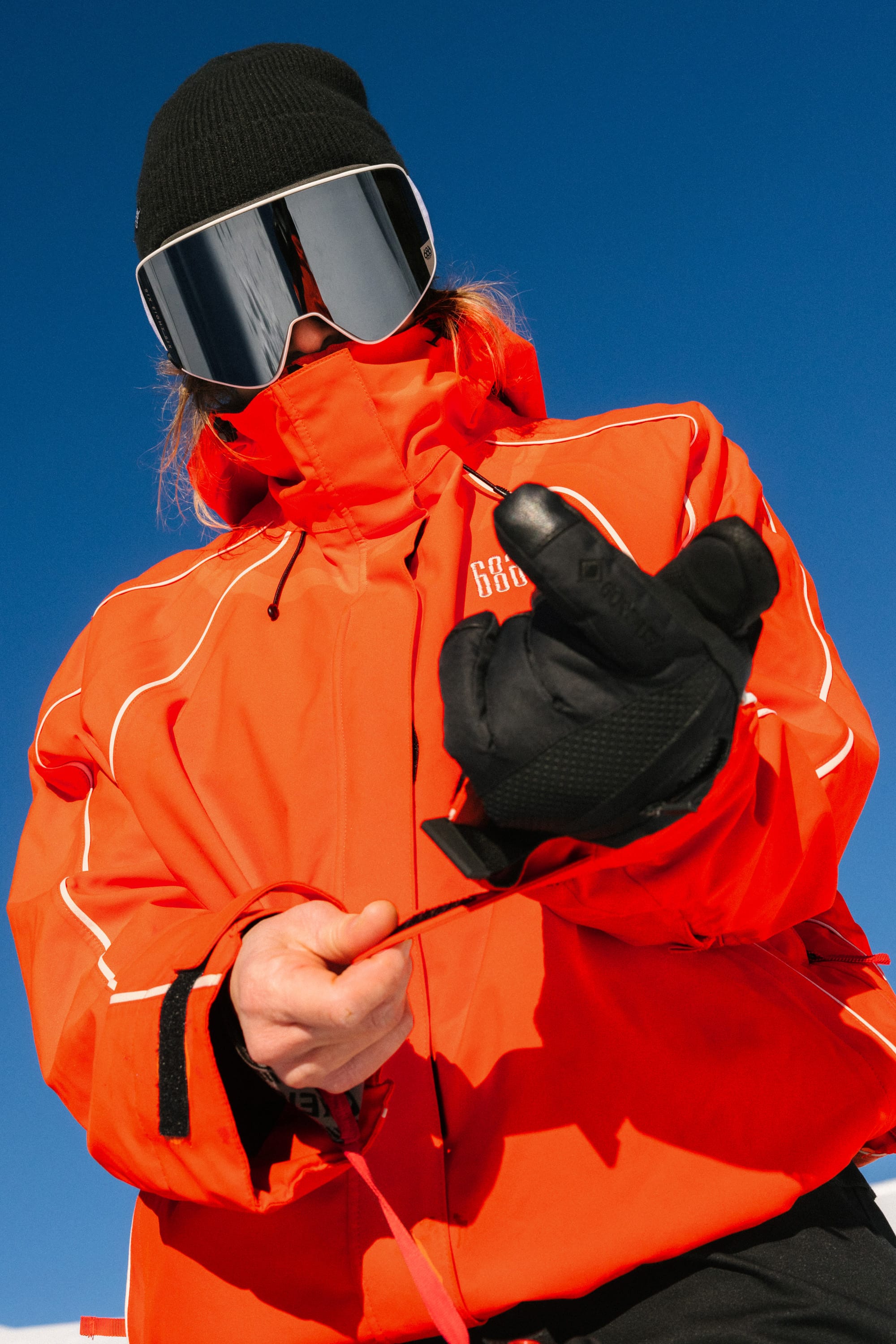 A person wearing the 686 Women's Hologram™ Shell Jacket in bright orange, along with black gloves, a black beanie, and reflective ski goggles, stands outdoors under a clear blue sky, adjusting their jacket.