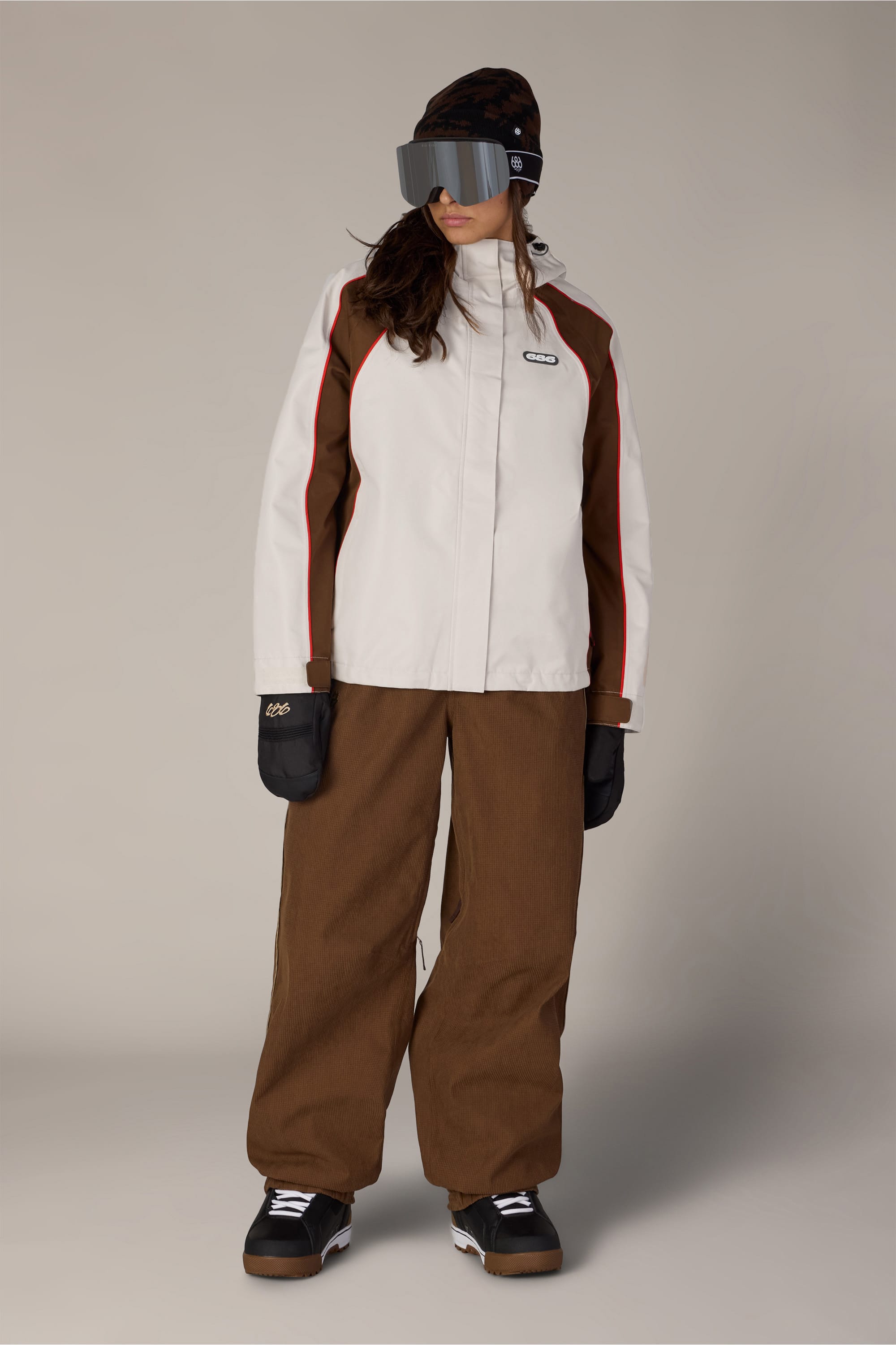 A person wearing brown pants, black gloves, black snow boots, a black beanie, large ski goggles, and the 686 Women’s Hologram™ Shell Jacket by 686 stands against a plain background.