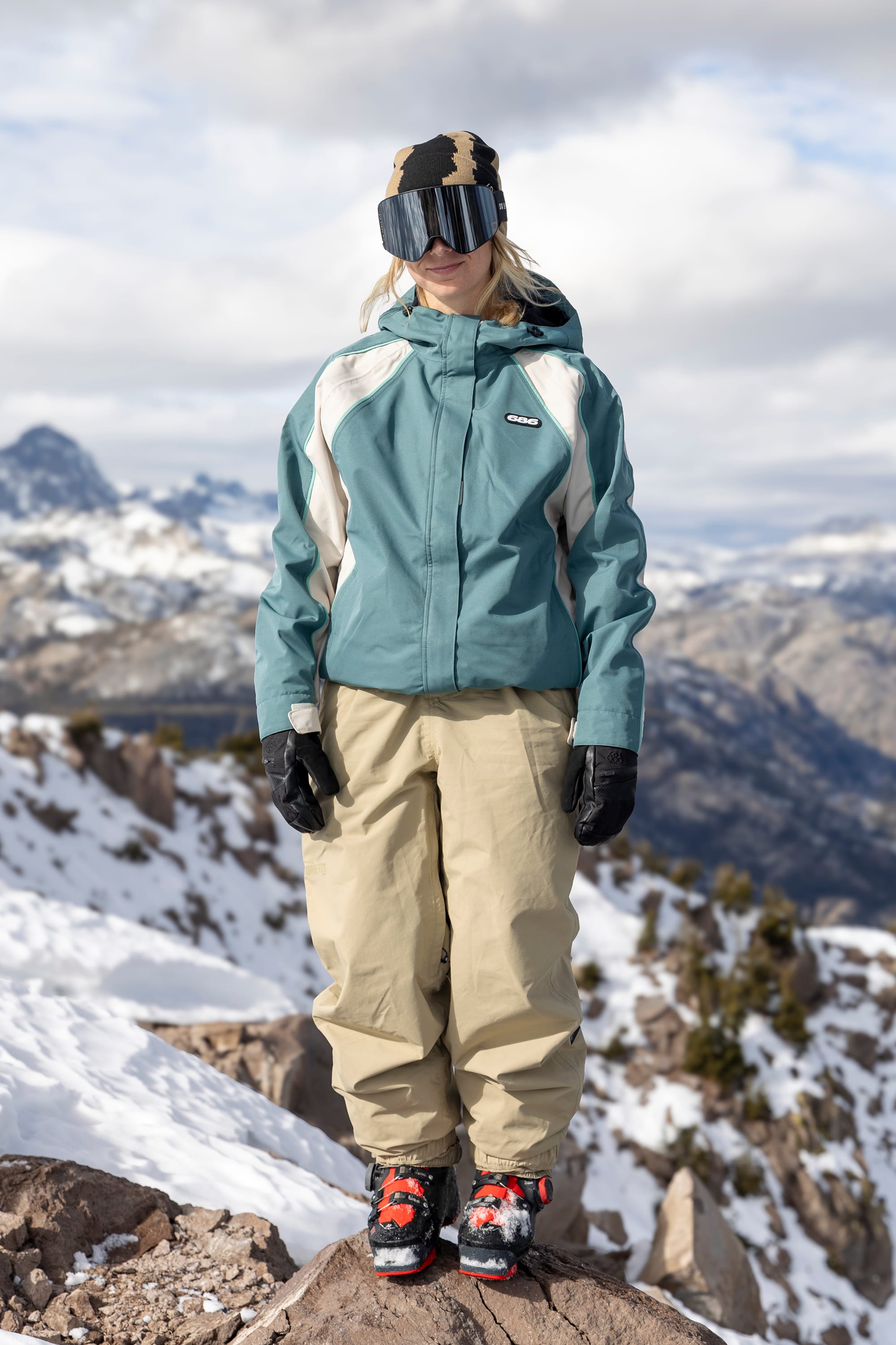 A person wearing the 686 Women’s Hologram™ Shell Jacket by 686, along with tan pants, black gloves, goggles, and boots, stands on a snowy mountain with a scenic view of snow-covered peaks in the background.