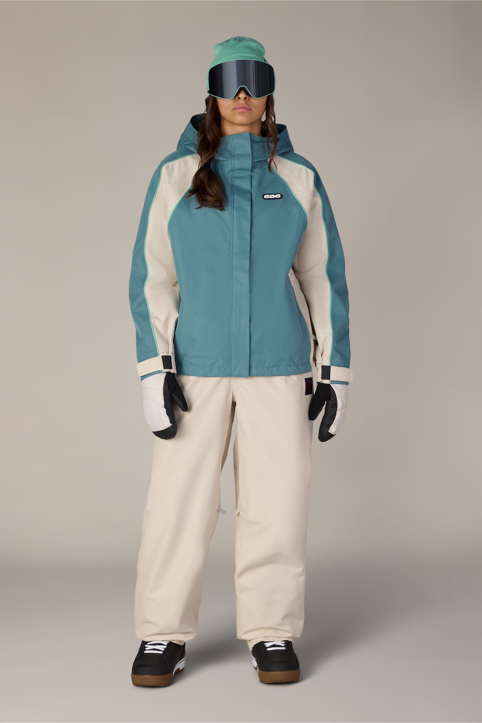 Wearing the 686 Women’s Hologram™ Shell Jacket by 686, black snow boots, white gloves, teal cap, cream ski pants, and large black ski goggles, the person stands against a plain light background.