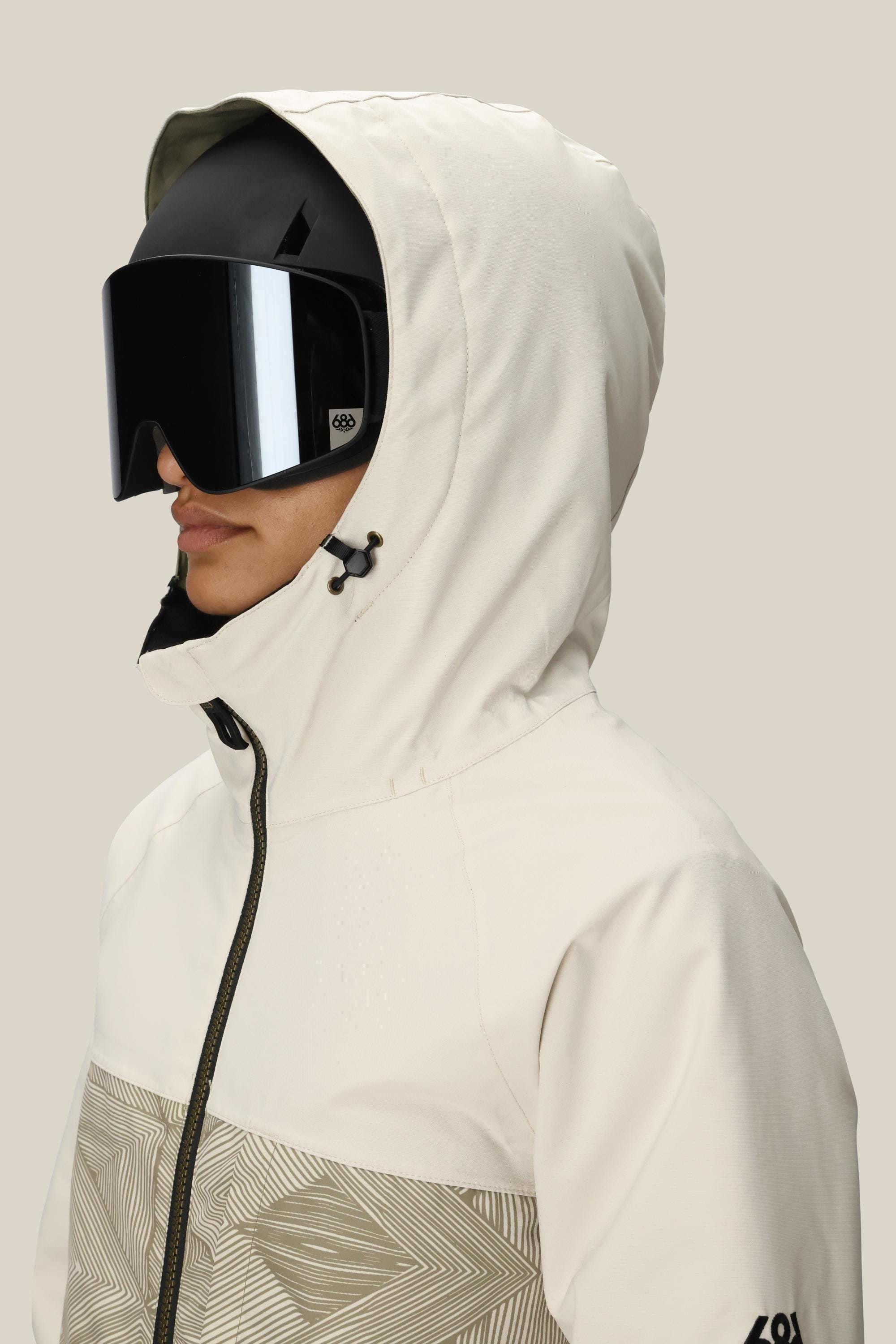 A woman wears the 686 Women's Athena™ Insulated Jacket in cream with a hood and large black ski goggles, facing slightly to the side against a plain light background.
