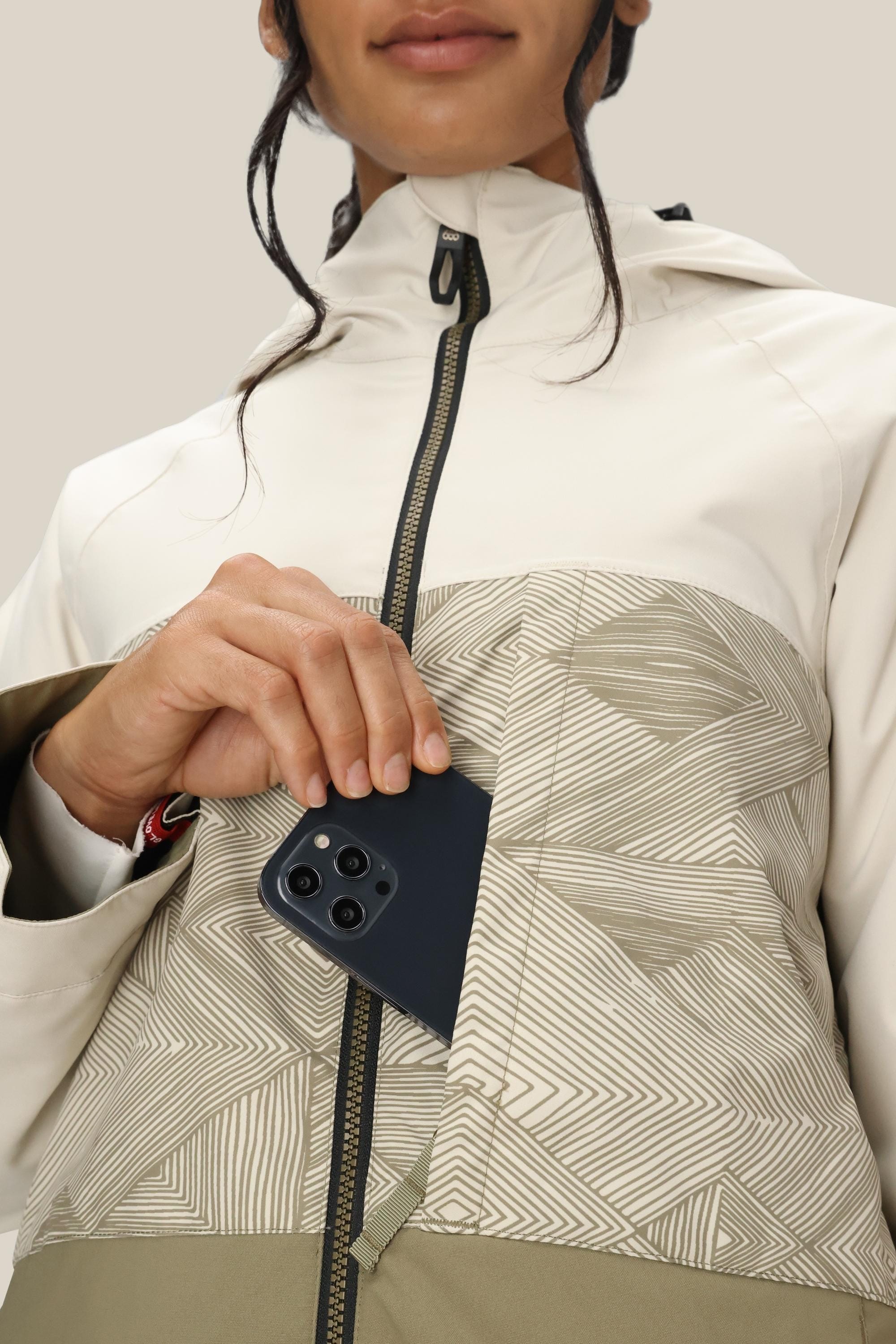 A woman in a light 686 Women's Athena™ Insulated Jacket puts a dark smartphone into the jacket’s front zip pocket. Her face is partly visible against a plain background.